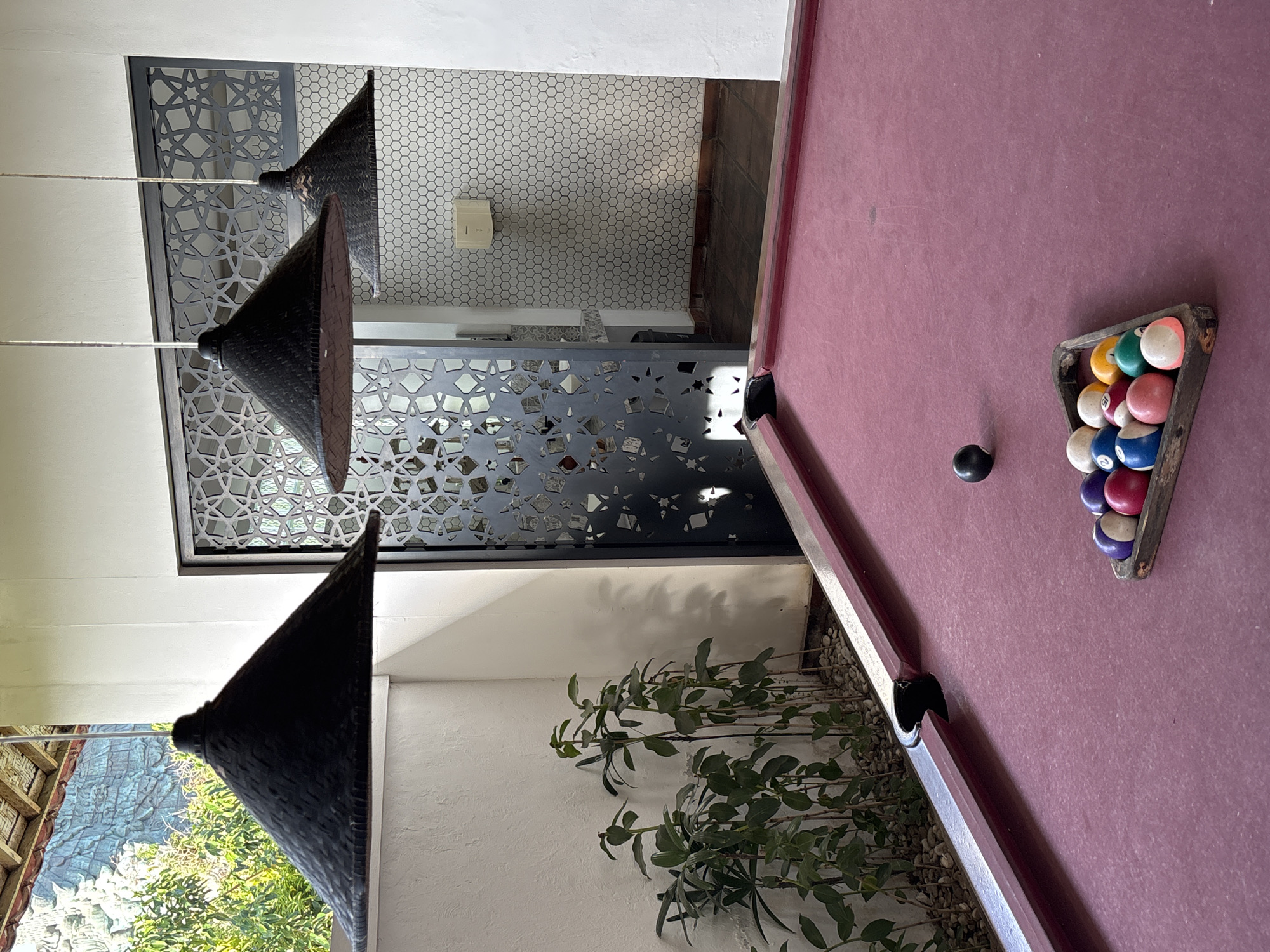 A slightly canted, interior medium shot shows a section of a pinkish-purple pool table with the racked balls on the right side, and a black cue ball sitting to the left of the racked balls. The table is lined in black. Behind the table is a wall with a row of bushes along the bottom of it. Above the wall are two large pendant lights, shaped like dark pyramids. Behind the lights is a dark grey, decorative metalwork screen that partially obscures the view of another area covered in small, white hexagonal tiles. The location is identified as Kuta Selatan, Indonesia.