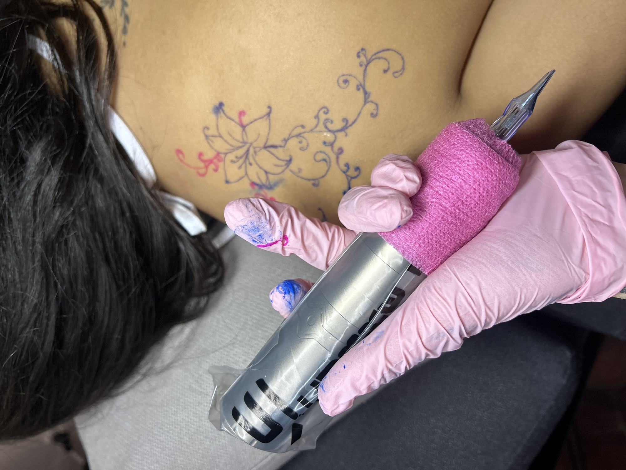 The image depicts a person receiving a tattoo on their upper back, presumably in a studio or private residence in Buenos Aires, Argentina. A tattoo artist, wearing pink protective gloves, is shown holding a tattoo machine wrapped in a clear plastic sleeve and with pink material around the grip. The tattoo design on the back features a large flower with blue outlines and pink shading, connected to a vine-like design with curly tendrils in blue. The client has dark hair that obscures the left side of the image. The background includes a grey-colored surface on which the client is lying and a dark grey surface where the tattoo artist is sitting.