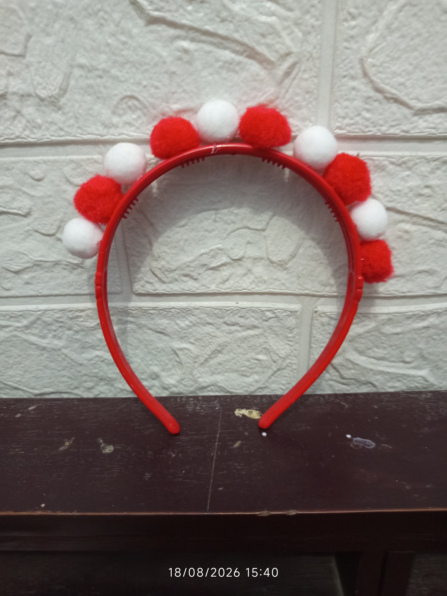 A red plastic headband with multiple red and white pom-poms is placed on a dark brown wooden surface. The headband is positioned horizontally, with the pom-poms distributed along its upper edge in an alternating pattern of red and white. The background is a white brick-patterned wall. The image is a close-up shot, with the timestamp "18/08/2026 15:40" visible in the lower right corner. The scene is indoors and appears to be a product photo or display.