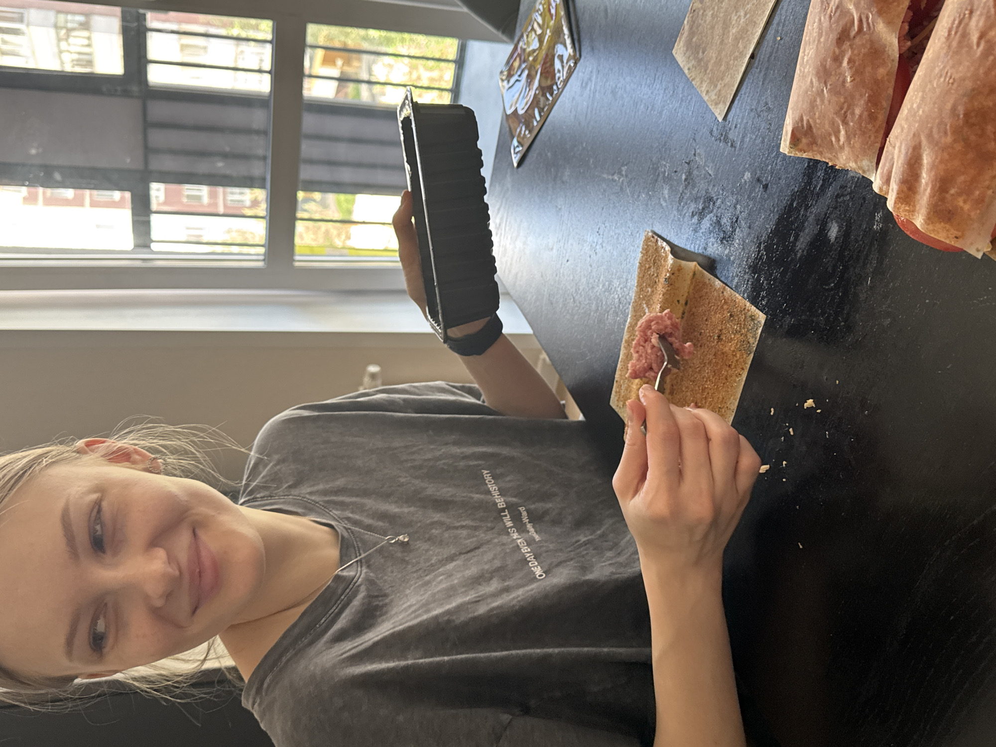 A person is preparing food at a table. The person is holding a black container in their left hand and using a fork in their right hand to spread a pink mixture onto a square piece of what appears to be a tortilla or lavash bread. There are several rolled-up, presumably finished food items to the right. Other items are on the table, including a tablet. The person is wearing a gray t-shirt and a black wristband. A window with blinds is visible in the background, revealing buildings outside. The location appears to be indoors. Without access to additional verifiable information, I can not confirm if the city is in Perm, Russia.