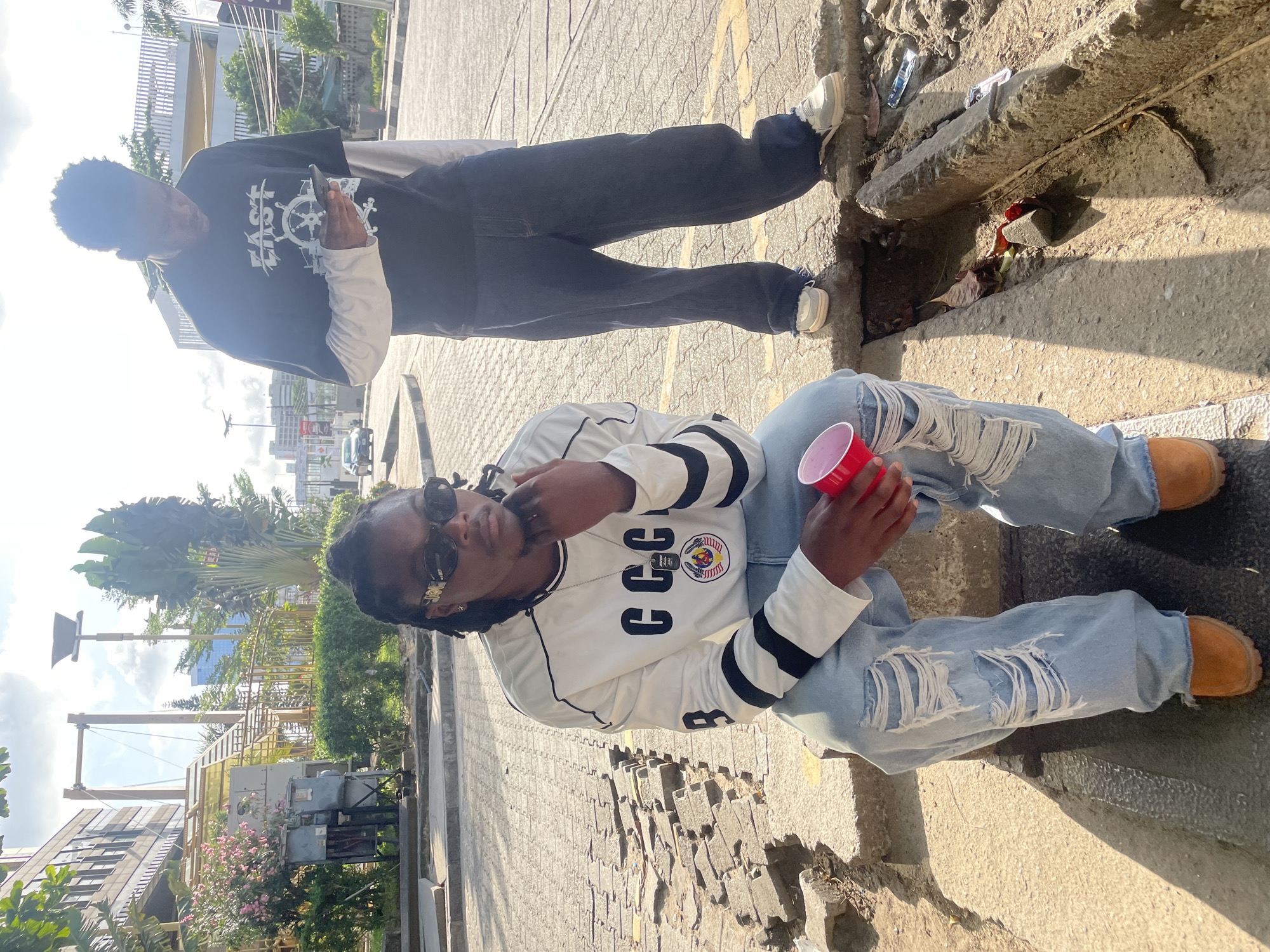 Two people are in an outdoor urban environment. One person is in the foreground, sitting on a ledge or low wall, wearing light blue ripped jeans, brown shoes, a long-sleeved white shirt with black stripes on the sleeves and "CCC" printed on the front, and sunglasses. They are holding a red plastic cup. The other person is standing behind them, partially out of focus, wearing dark jeans, a long-sleeved black shirt with a white graphic on the front, and white shoes. The ground appears to be paved with patterned stones or tiles. In the background, there is greenery, buildings, utility equipment, and a clear sky with some clouds.