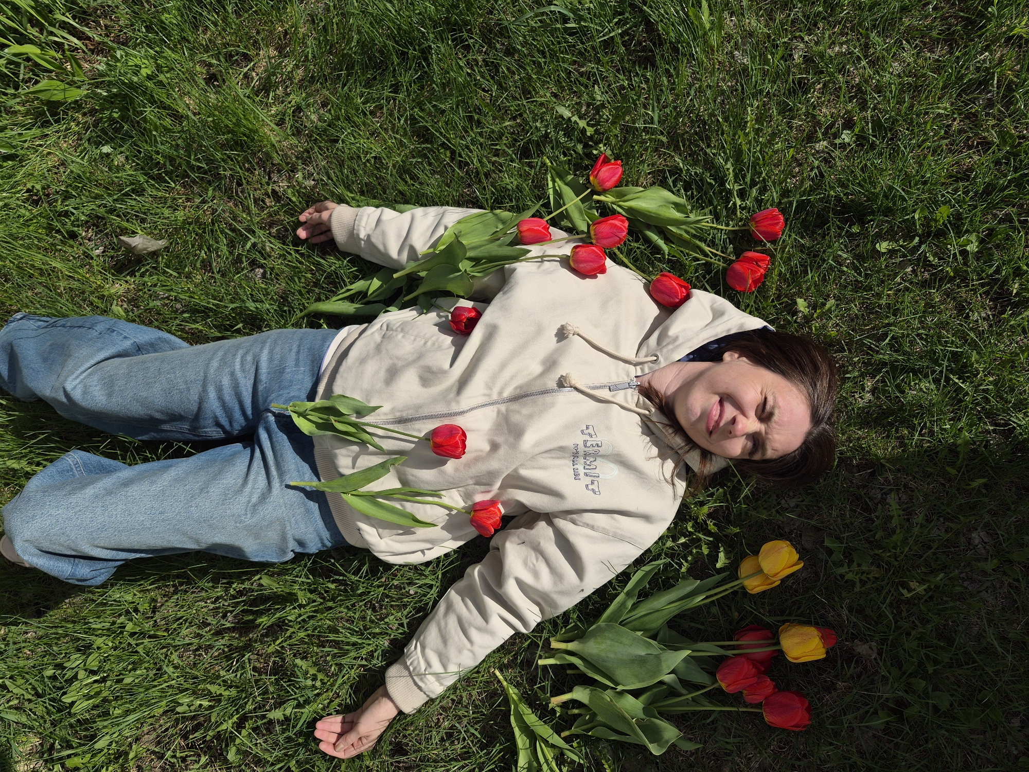 A young woman lies on her back in green grass, possibly in a park or lawn. She is wearing blue jeans and a light-colored zip-up hoodie. Red and yellow tulips are scattered around her body. The photo appears to be taken from an overhead perspective. The city of Chaikovsky, Russia is not evident in the image.