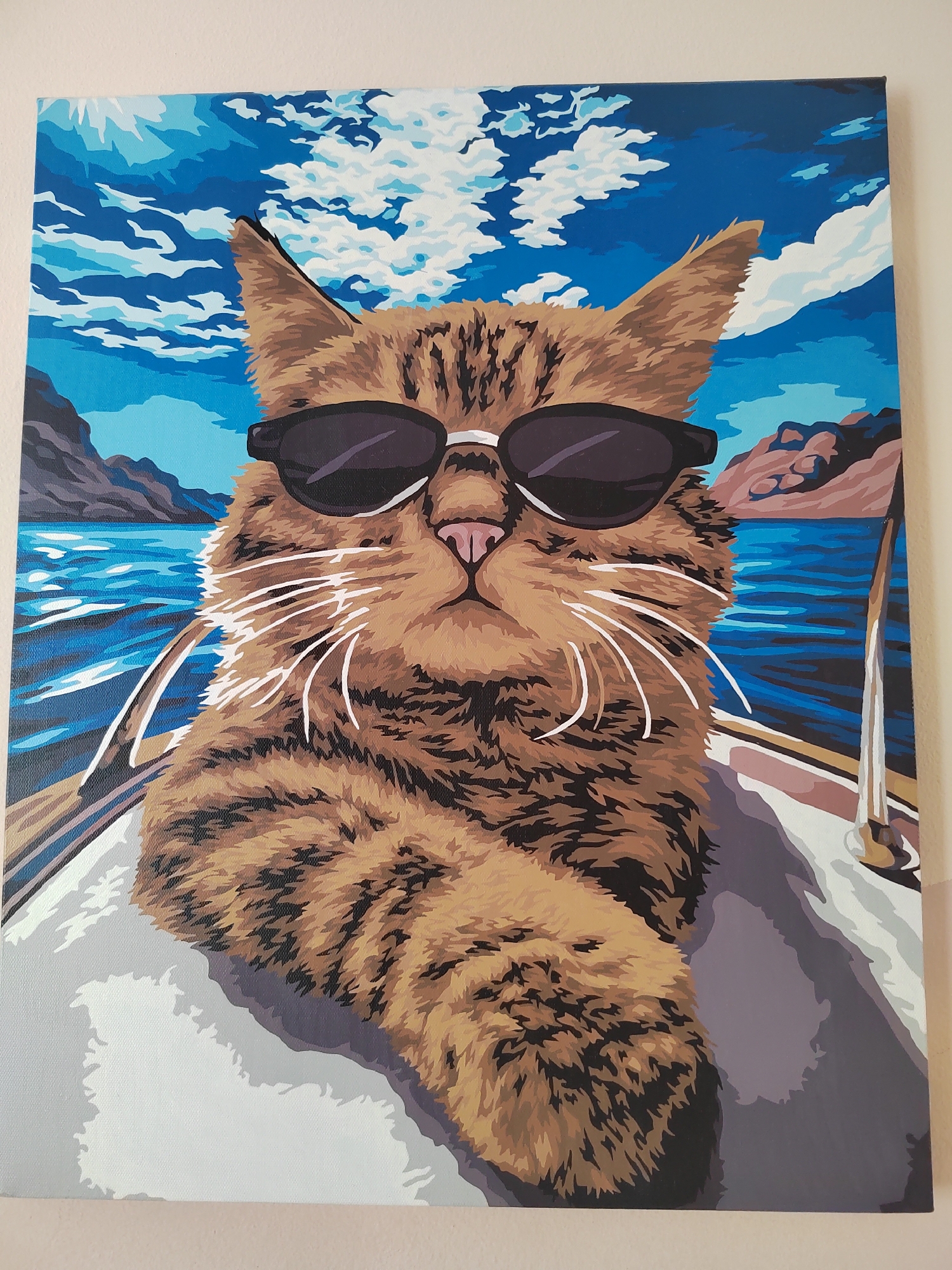 The image is a painting of a tabby cat wearing sunglasses, sitting on a boat. The cat is in the foreground, looking directly at the viewer. The background depicts a body of water, mountains, and a blue sky with clouds. The painting is done in a stylized, graphic manner with bold colors and defined shapes. There are no people or specific Vityazevo landmarks visible in the painting.