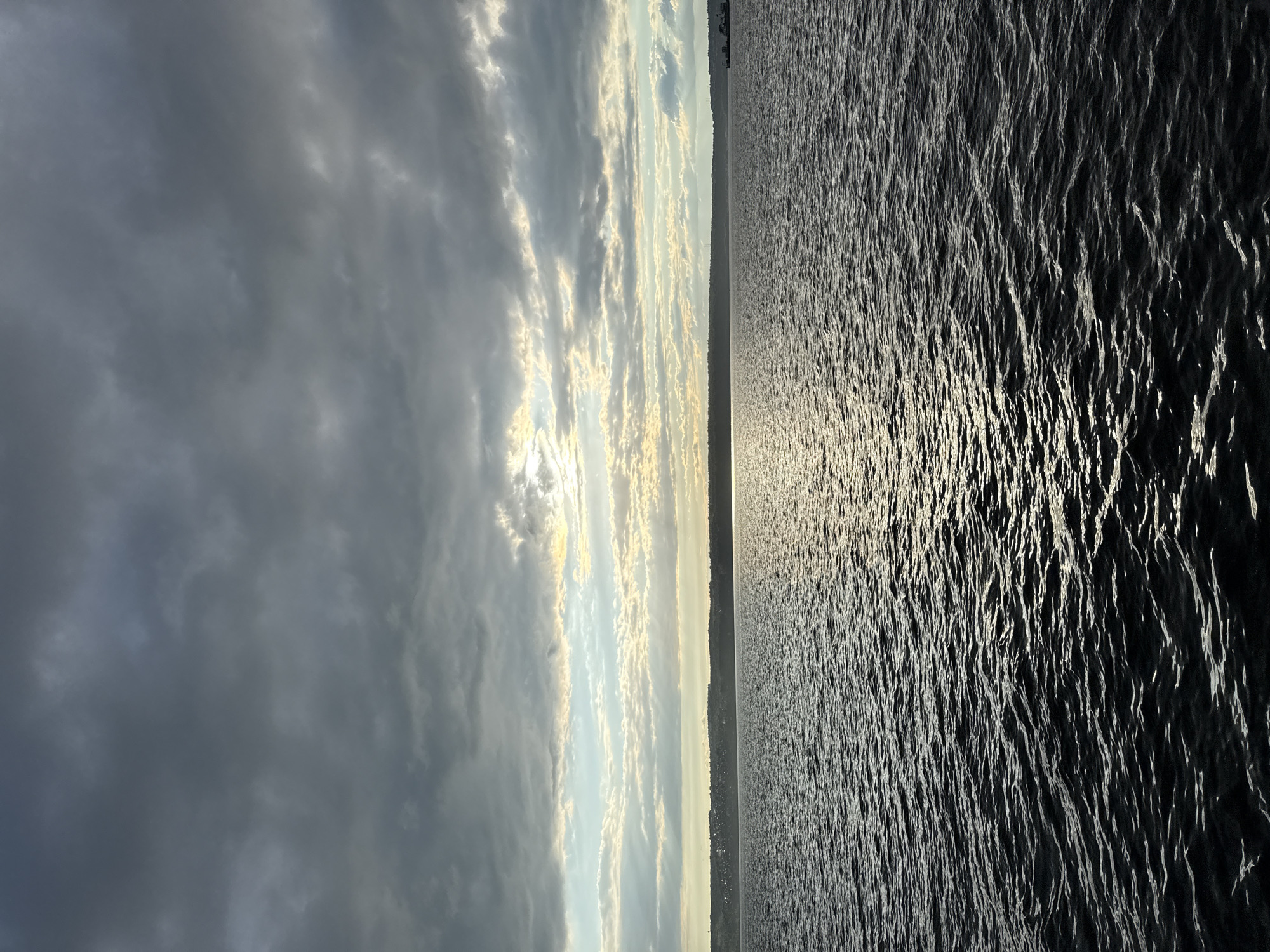 The image depicts a wide body of water, likely a river or lake, under a dramatic, cloudy sky. The sun is partially visible through the clouds, casting a shimmering reflection on the water's surface. The horizon line is marked by a low-lying landmass, possibly the shore of Kazan, Russia, with a faint line of buildings and trees visible. A small boat or barge is present on the right side of the horizon. The water is dark and choppy, with ripples creating a textured appearance. The clouds are a mixture of dark grey and lighter, illuminated patches, suggesting an overcast but potentially clearing sky. The overall mood is serene and slightly dramatic due to the lighting and cloud cover.