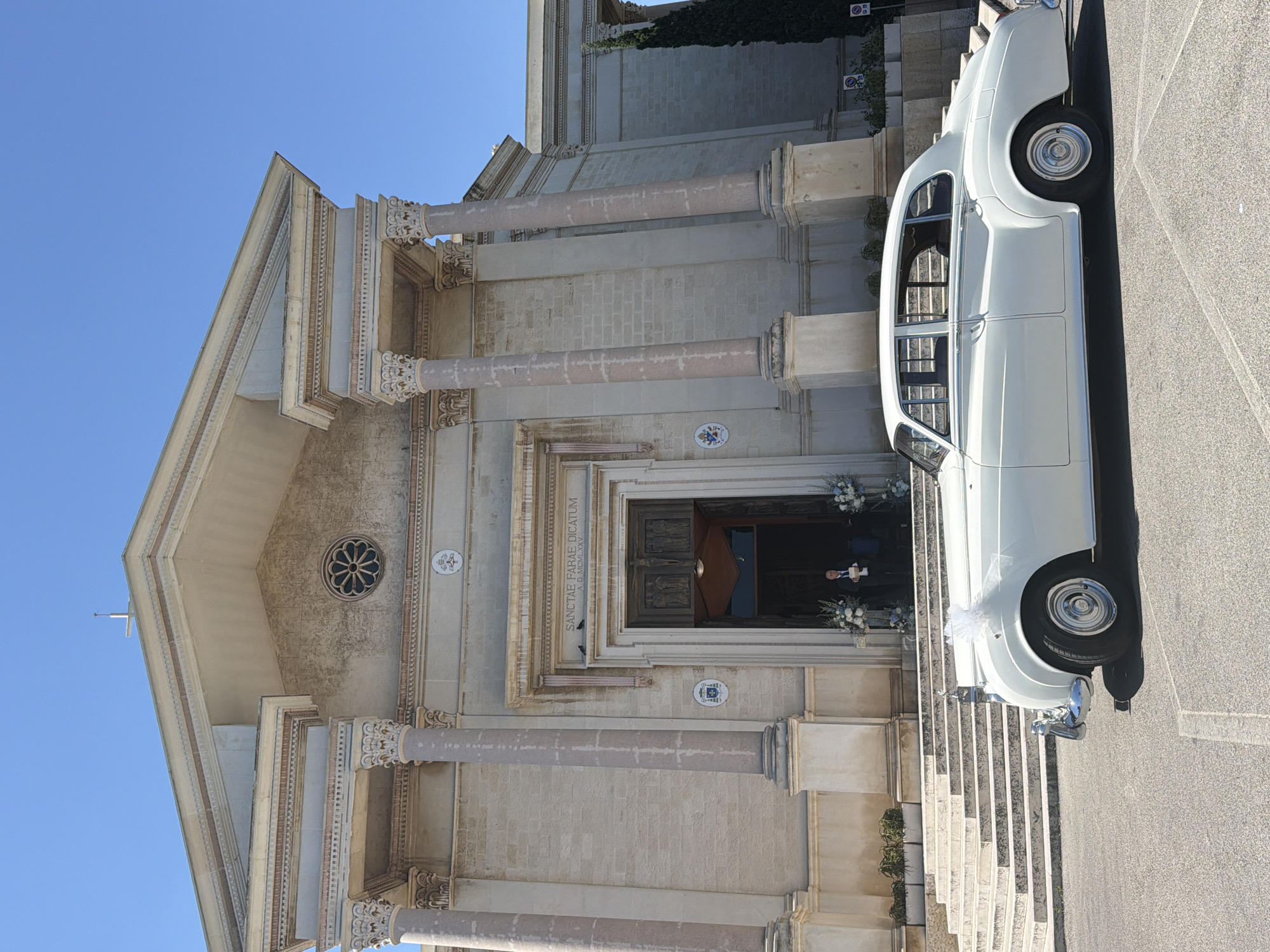 The image shows a white vintage Rolls-Royce parked on a paved surface in front of a large, light-colored stone building, likely a church or a significant public building, with grand columns and a triangular pediment. The building has a prominent entrance with a carved wooden door and is adorned with inscriptions and emblems. A man in a suit is standing in the doorway, holding a piece of paper. There are floral arrangements on either side of the entrance. To the left of the entrance, there are wide stone steps leading up to the building. To the right, a tall, dark green evergreen tree is visible. The sky is a clear, bright blue. The car is decorated with a white ribbon tied on the hood. Based on the architectural style and the presence of a church, it is plausible this scene is in a historical European city. Given the specific context of Bari, Italy, this building is likely a church within the city.