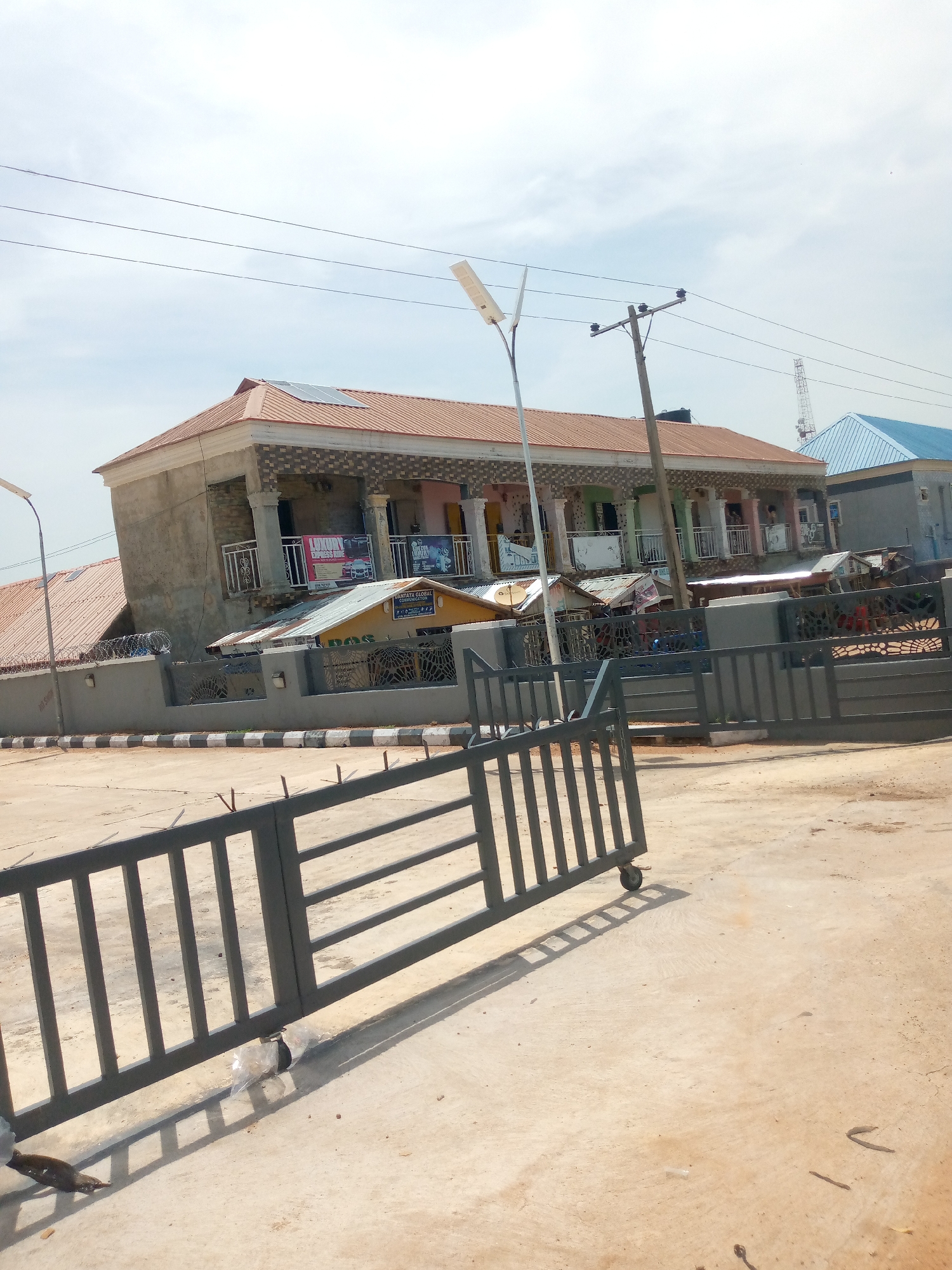 The image shows a building with a multi-colored tiled roof, a balcony with several pillars, and windows on the upper floor. Solar panels are visible on the roof. Below the balcony, there are several shops or stalls with metal roofs. A grey metal gate with vertical bars and a circular design pattern is in the foreground, partially blocking the view of the building. A street light is positioned to the left of the building. In the background, there is a tall communication mast and another building with a blue roof. The ground in front of the gate is a light-colored concrete or paved surface. The scene appears to be in an outdoor setting in a city, likely during the daytime with a cloudy sky. The context provided mentions Jalingo, Nigeria, suggesting this is a location within that city. There are no visible people in the foreground or clearly discernible in the building's windows or at the stalls. The overall impression is one of a commercial or mixed-use development.