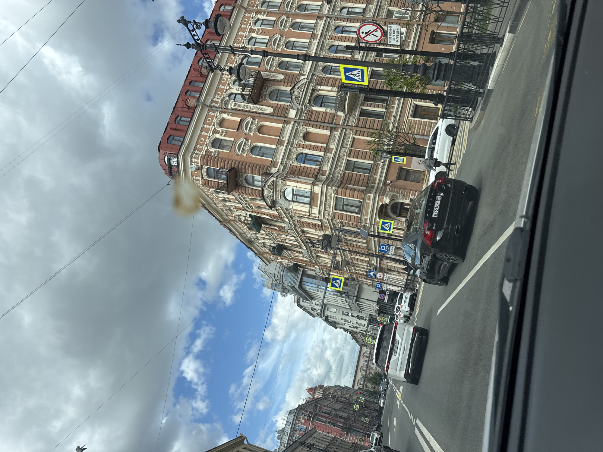 Captured from within a vehicle, the image depicts a street in Saint Petersburg, Russia. The architecture is characterized by ornate, multi-story buildings with red roofs and detailed facades. The street is populated with cars, and pedestrian crossings marked by yellow and blue signs are visible. Power lines stretch across the cloudy sky, which shows patches of blue. The scene is urban, with a mix of old architectural styles and modern traffic.