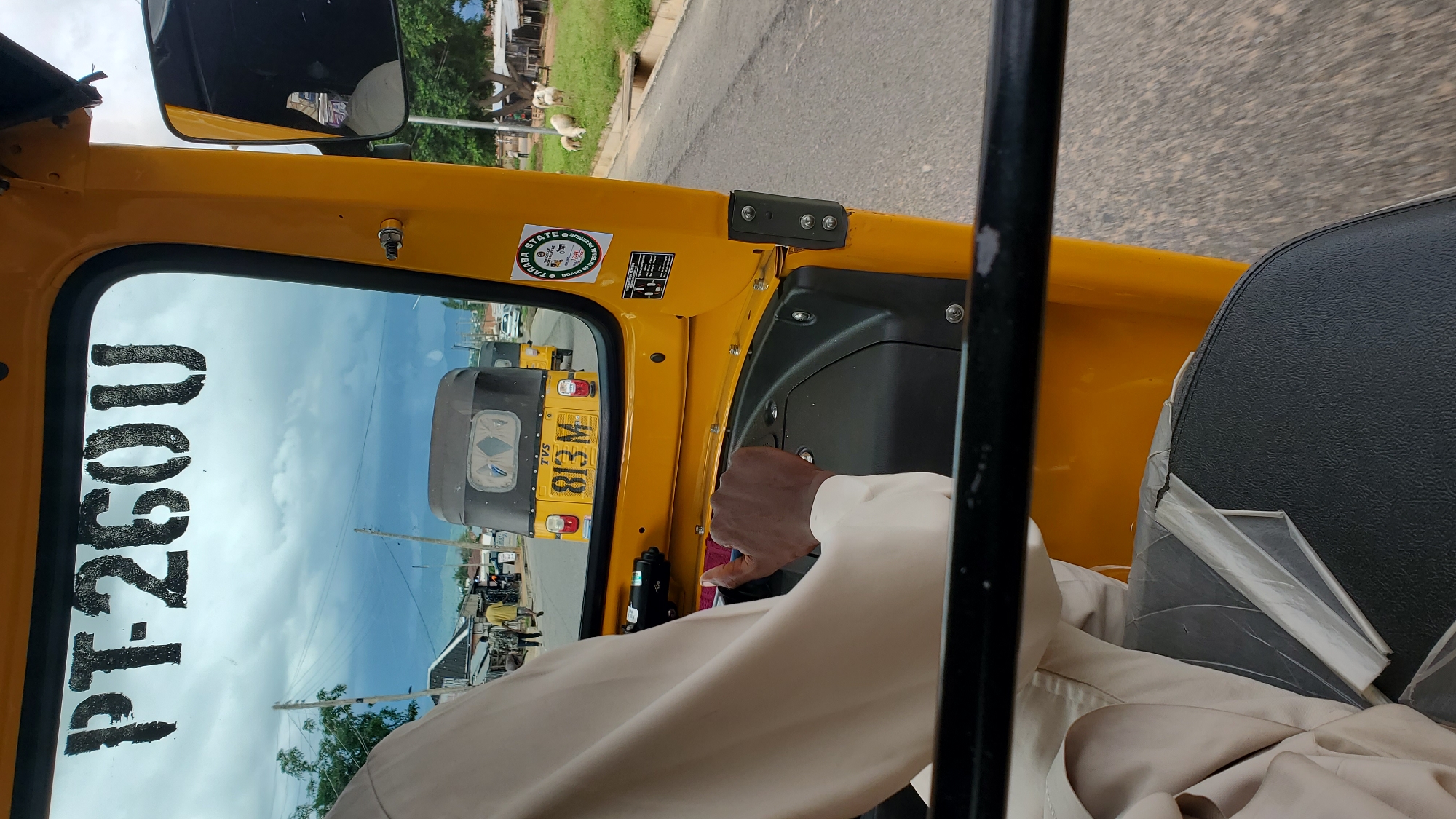 The image is a close-up, interior shot from the driver's perspective of a yellow auto-rickshaw in Jalingo, Nigeria. The windshield displays the registration number "PT-260U" stenciled in black. Through the windshield, other vehicles, including auto-rickshaws with visible license plates, are seen on a paved road. A sticker on the right side of the auto-rickshaw reads "TARABA STATE" with a logo. The driver's hand, holding the steering wheel, is visible in the foreground. In the background, there are buildings, trees, and grazing sheep, suggesting a semi-urban or rural setting within Jalingo. The sky is overcast with dark clouds.
