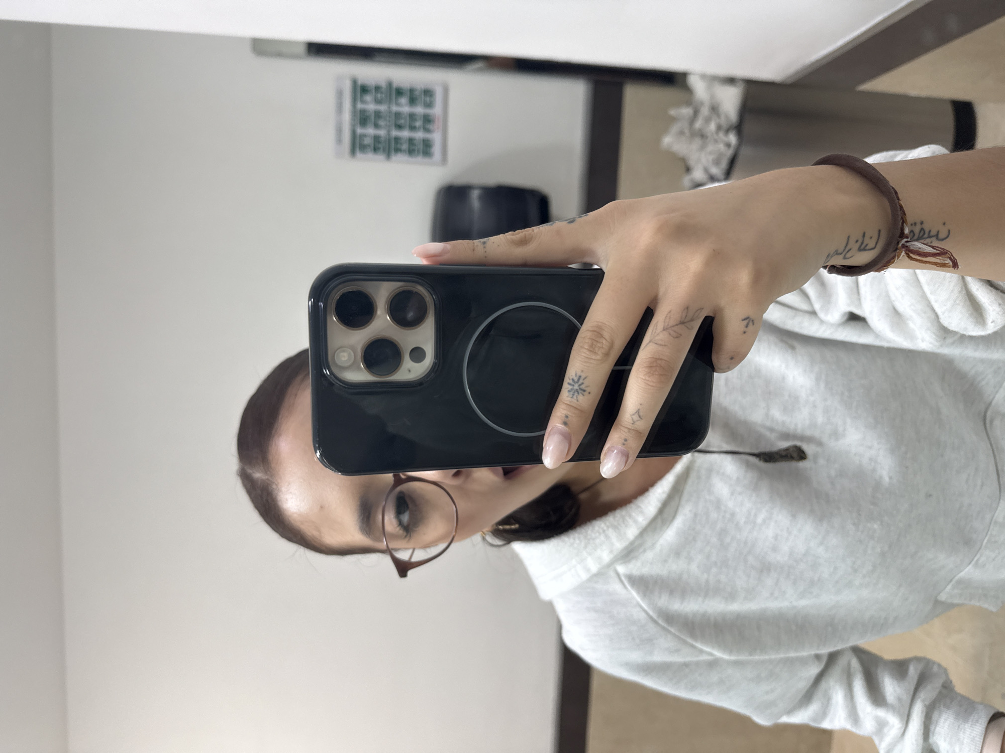 A person is captured taking a mirror selfie indoors, holding a black smartphone with a visible MagSafe ring on its case. The phone's camera module, featuring multiple lenses, is clearly visible. The person's face is partially reflected in the mirror, showing dark hair parted down the middle and wearing brown-framed glasses. Only the right side of their face is visible, with eyes looking towards the phone.
The individual is wearing a light-colored, possibly white or light grey, hooded sweatshirt. Their hands and forearms are prominent, revealing several small tattoos. On the fingers, there are delicate designs including a leaf branch, a snowflake-like symbol, and a small star or cross. A tattoo of Arabic script is visible on the inner right forearm, partially obscured by a brown elastic hair tie worn as a bracelet. The fingernails have a natural, light-colored polish.
The background reflected in the mirror consists of a plain white wall with a dark object (possibly a chair or bag) in the mid-ground. Above this, a rectangular sign or poster with a grid-like pattern of illegible green text or symbols is affixed to the wall. The lighting appears to be ambient indoor light. The setting is an indoor space, potentially a room or hallway, in San Andrés Cholula, Mexico.