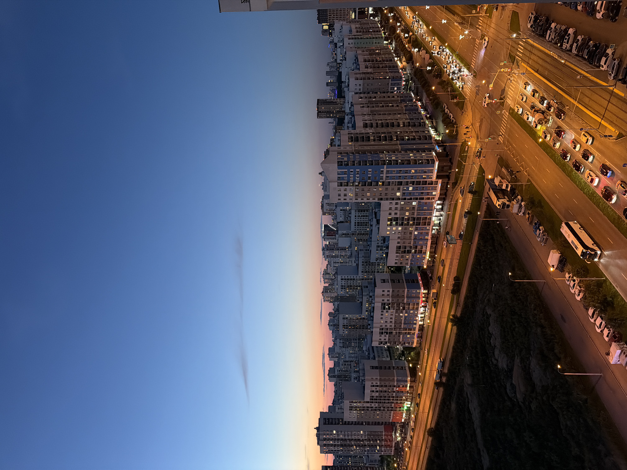 The image depicts a city skyline at dusk in Yekaterinburg, Russia. The sky transitions from a deep blue at the top to a soft orange and pink hue near the horizon, indicating the setting sun. Numerous residential buildings, characterized by their multi-story architecture and uniform design, dominate the mid-ground. Many windows are illuminated, suggesting occupancy.
A multi-lane road runs through the foreground and mid-ground, with a steady stream of vehicles visible. Headlights and taillights illuminate the asphalt, creating streaks of light. Several cars, trucks, and a bus are identifiable. Pedestrians are sparsely seen on sidewalks and at crosswalks. Streetlights cast a warm glow on the roadways and sidewalks. Adjacent to the road, there is a patch of dark, uncultivated land with some vegetation. The overall impression is that of a modern, active urban environment transitioning from day to night.