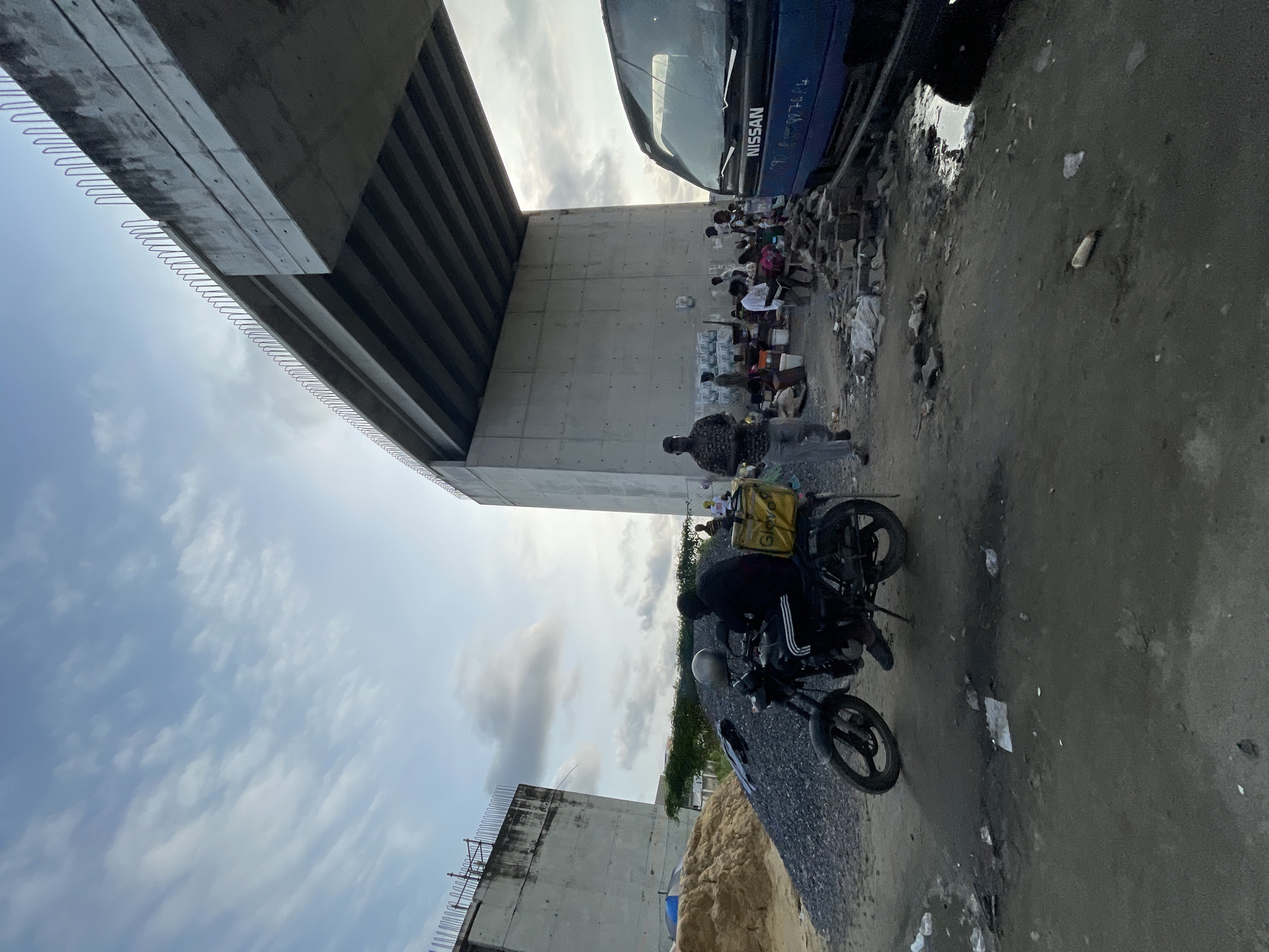 Underneath an elevated concrete highway construction in Lekki, Nigeria, a group of people are gathered. A blue Nissan truck is parked on the right side of the frame. To the left of the truck, a small group of individuals are seated around what appears to be a makeshift market or gathering area, with various objects and containers scattered around. Further left, a motorcycle with a yellow delivery box is parked, and a person in black clothing is seated on it. In the background, the massive concrete pillars and beams of the unfinished highway dominate the scene. The ground is uneven and dusty, with piles of gravel and sand visible on the left. The sky above is partly cloudy.