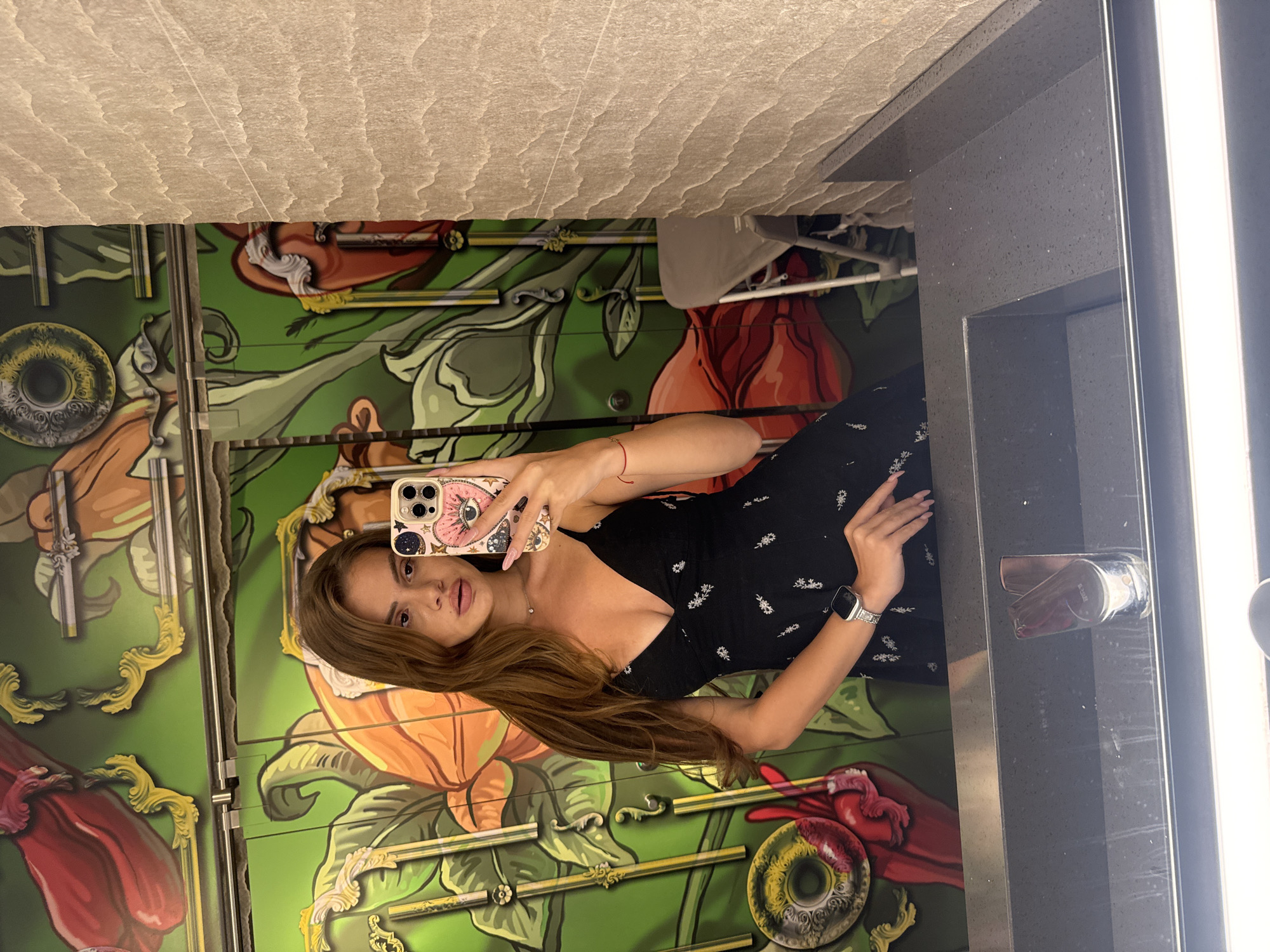A young woman with long, brown hair takes a mirror selfie in a restroom. She is wearing a black dress with a plunging neckline and a delicate floral pattern. Her right hand is raised, holding a phone with a pink case adorned with a celestial eye motif. On her left wrist, she wears a silver smartwatch. The background features a vibrant mural depicting large, stylized flowers and ornate golden elements on a green background. To the right of the mural, a beige, textured wall is visible. A grey countertop with a sink is in the foreground. The restroom appears to be in a modern establishment, possibly a restaurant or bar, located in Plovdiv, Bulgaria.