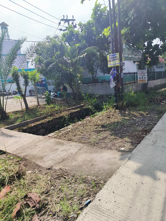 The image shows a paved walkway in Cileunyi, Indonesia, located at coordinates -6.947, 107.731. The walkway runs along a drainage ditch lined with stone blocks. On the left, there are trees and bushes, with a partially visible building in the background. A sign with the words "TERAPI SARAF KEJEPIT" is posted on a utility pole along the side of the walkway. There is a blue fabric hanging from the pole. The pavement is cracked and uneven, with signs of dirt and debris. There is a large tree on the left side of the image, and a smaller tree in the middle. The image was taken during the day in sunny conditions. The ground is dry and dusty, with a few areas of grass and weeds. The walkway is empty of people.