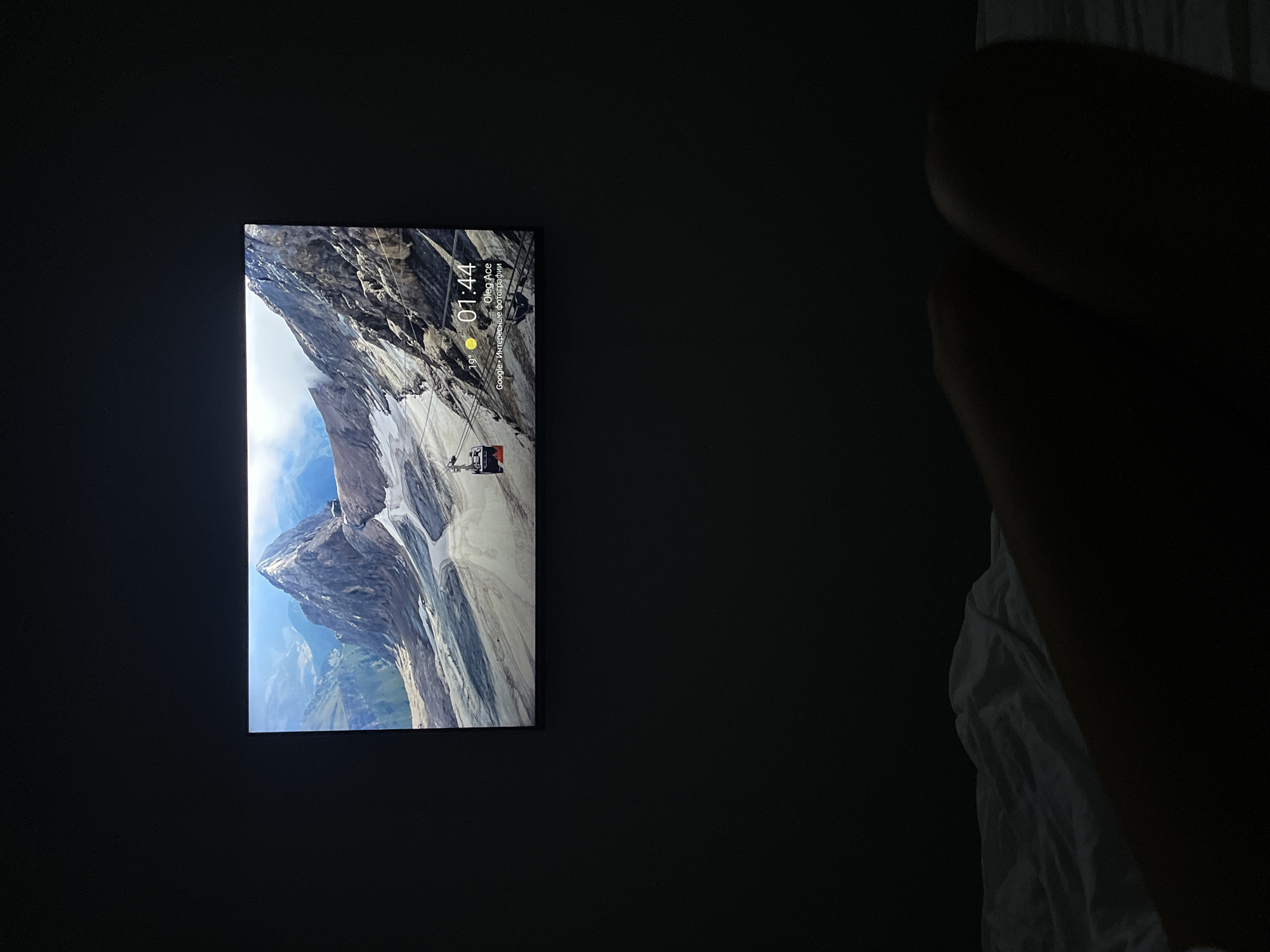 An indoor scene, situated in Baranovka, Russia, depicts a dimly lit room illuminated primarily by a television screen mounted on a dark, plain wall. The screen displays a high-resolution image of a snow-capped mountain range, featuring a prominent, jagged peak and an extensive glacier. A red cable car cabin is visible on the lower left of the screen, traversing along a cable system against the mountainous backdrop. Overlayed text on the screen indicates a temperature of 19 degrees with a sun icon, the time 01:44, and credits "Oleg Ace" and "Google • Интересные фотографии" (Google • Interesting photos). In the foreground, the lower legs of an individual are visible, resting on wrinkled white bedding, suggesting a person lying down and viewing the screen. The overall ambiance is dark, with the television serving as the sole significant light source.