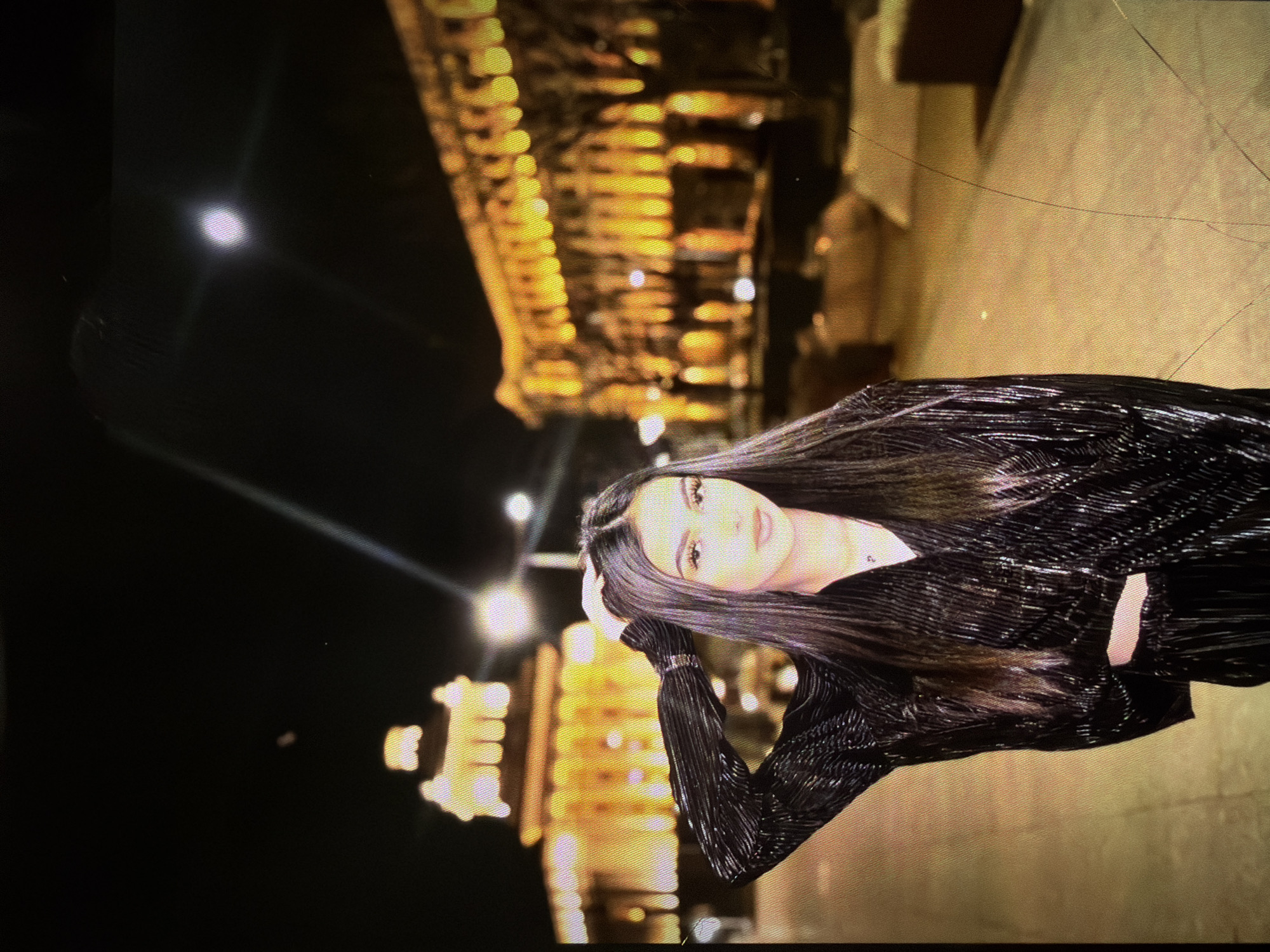 A young woman with long, dark hair is standing outdoors at night, posing for a photo with her right hand on her head. She is wearing a black, sparkly outfit. In the background, illuminated buildings with golden lights are visible, suggesting an urban setting. The sky is dark, and there is a bright light, possibly the moon or a streetlamp, in the upper right portion of the image. The ground appears to be paved.