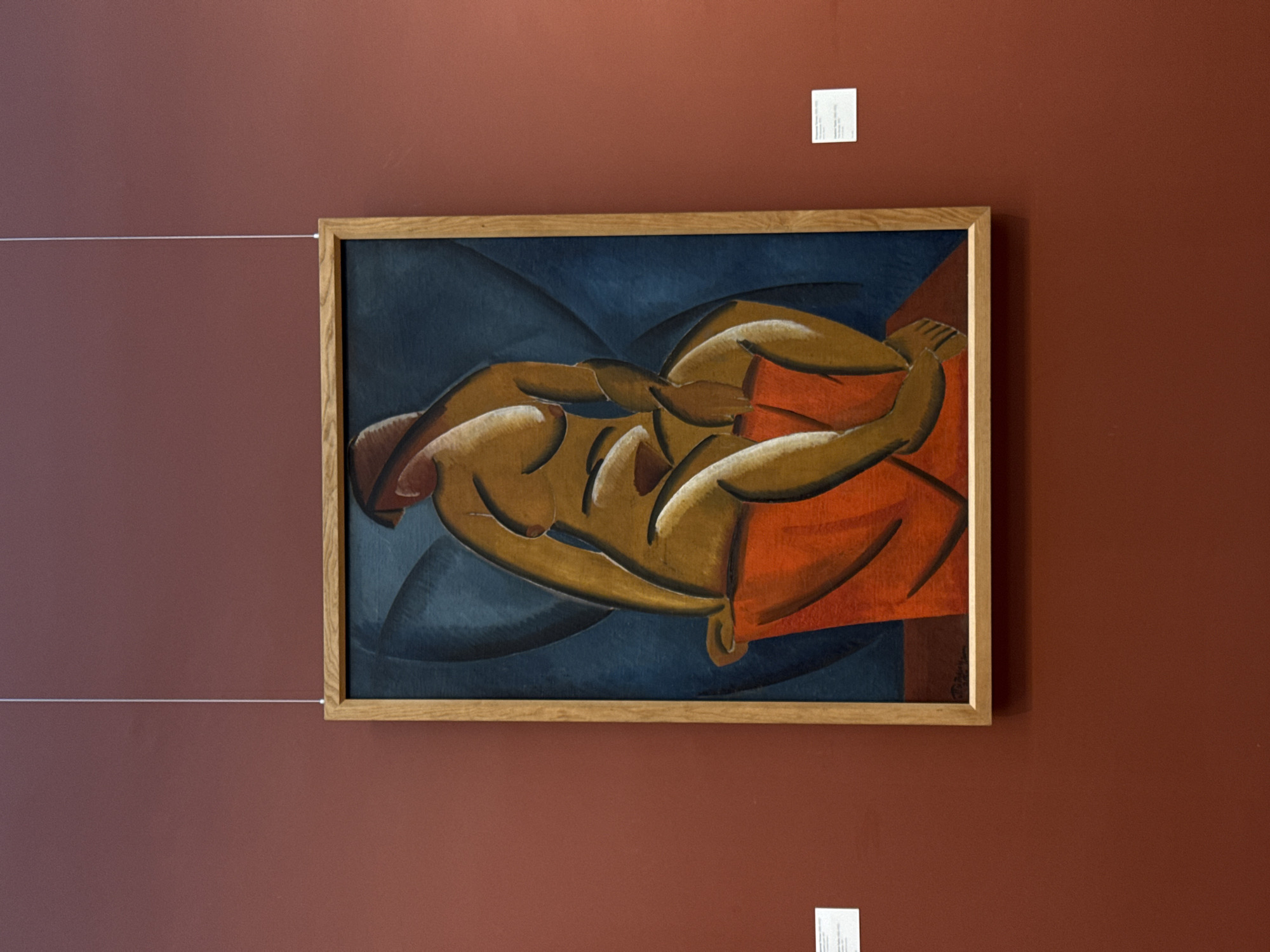 A framed painting of a nude or partially nude figure in shades of brown and orange against a dark blue background hangs on a brown wall. The painting is hung with a thin white wire. Above and below the painting are small white placards with printed text. The image appears to be taken indoors, potentially in a gallery or museum, based on the wall color and presence of descriptive placards. The city of Moscow, Russia is not apparent from this image.