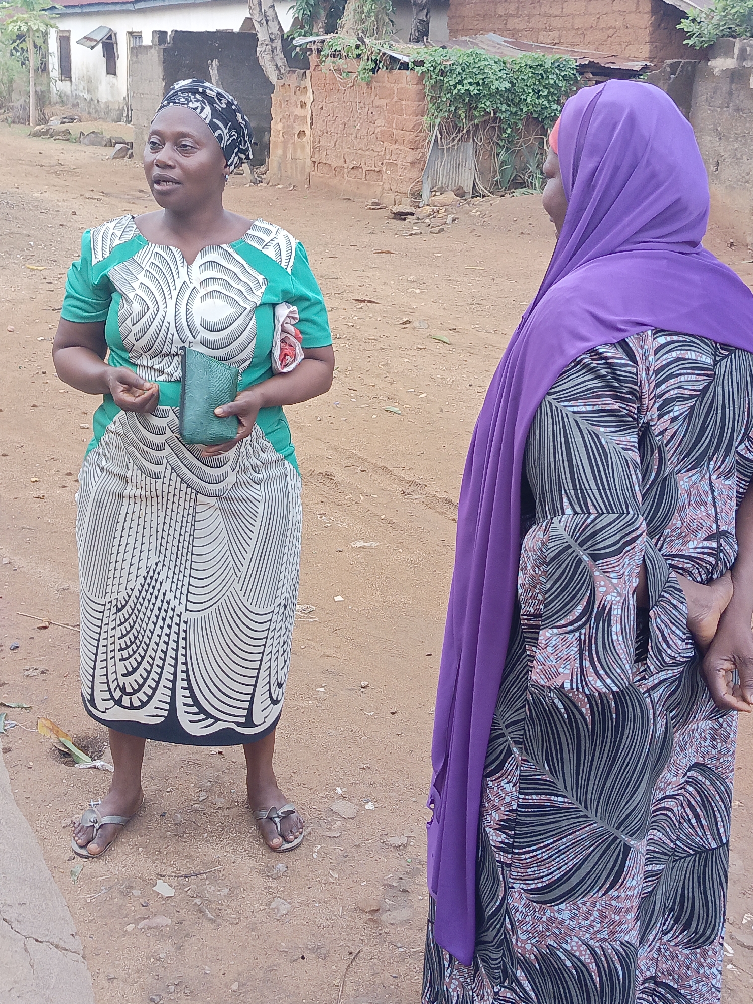 Two women stand on a dirt road in Akwanga, Nigeria. The woman on the left faces forward, wearing a green and white patterned dress, a black and white headscarf, and silver sandals. She holds a green clutch purse in her hands. The woman on the right is partially visible, wearing a patterned dress and a purple hijab. The background includes buildings constructed of brick and concrete and some foliage.