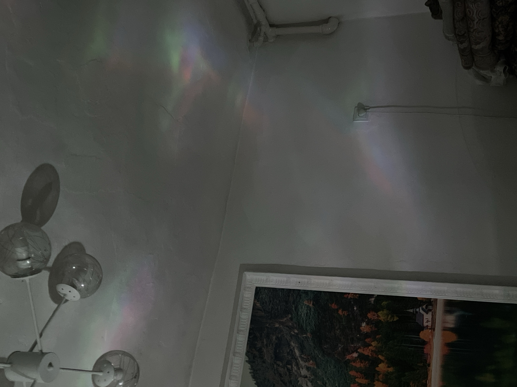 The image shows a ceiling fan with glass globe fixtures, casting colorful light reflections on a white, textured ceiling. The light patterns appear as streaks and splotches of pink, green, and blue. On the wall below, a landscape painting is partially visible, depicting a lake, trees with autumn foliage, and a small church. A white electrical outlet with a plugged-in cord is also visible on the wall. In the corner of the room, a stack of folded fabrics or blankets is present. The overall scene suggests an indoor residential setting. The location is not specified in Bishkek City, Kyrgyzstan.