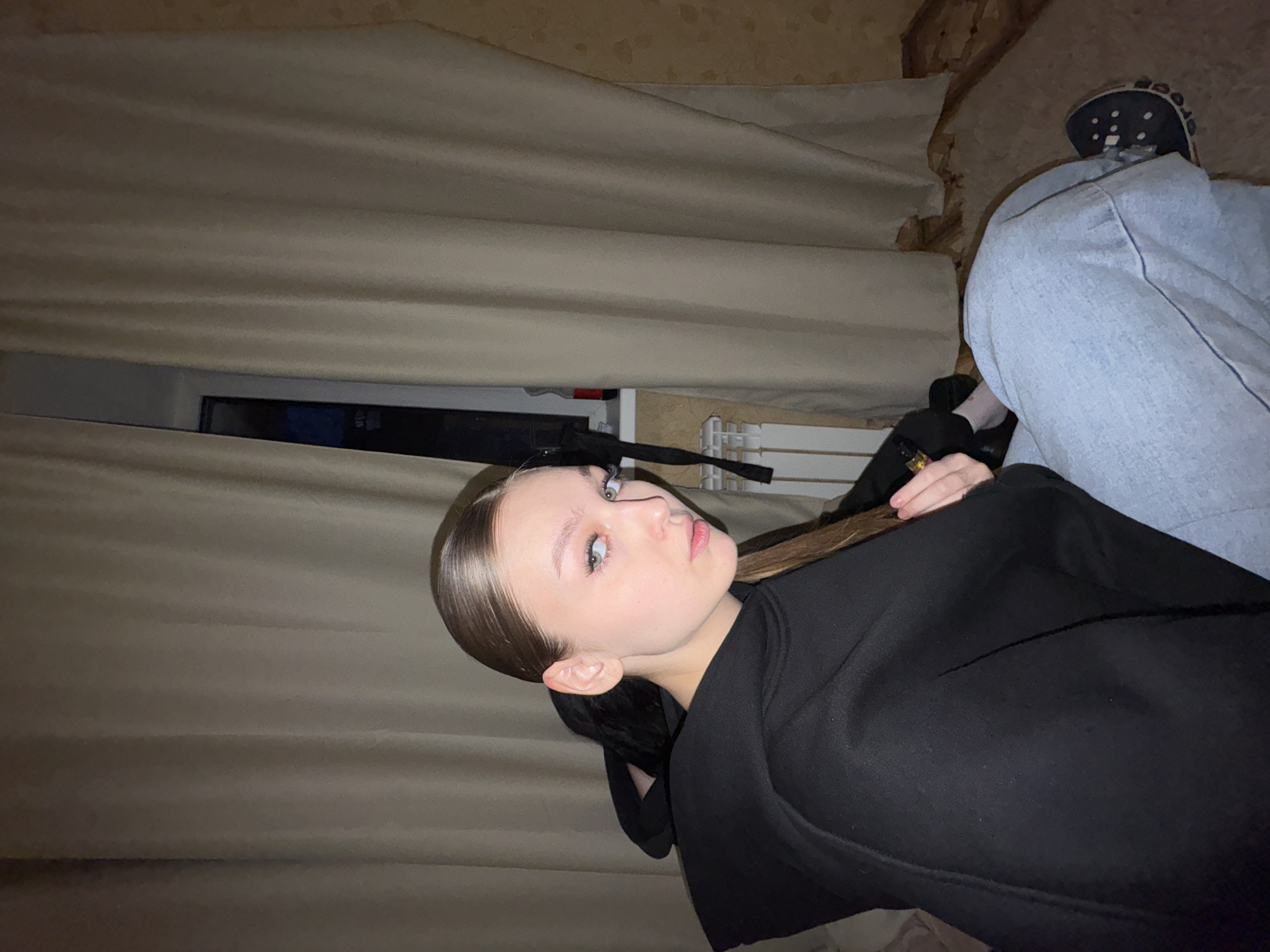 The image captures a young person in an indoor setting, likely a home in Chaikovsky, Russia. The main subject, a young female with light eyes and dark brown hair slicked back, is shown from the chest up, looking towards the camera with a neutral expression. She is wearing a black top or hoodie. In her left hand, she holds a small, golden-colored object, possibly a vape pen.
The background reveals light beige or light brown curtains that appear drawn, partially obscuring a window to the left. A glimpse of a white radiator or heater is visible below the window on the right side of the main subject. The ceiling above the curtains has a textured, yellowish-brown appearance.
In the upper right corner of the image, parts of another person are visible, specifically a leg clad in light-colored (possibly light blue) jeans and a dark blue Croc shoe. This person appears to be seated or reclining, parallel to the main subject. The overall lighting is dim, suggesting the photograph was taken in the evening or at night. No discernible text is visible in the image. The scene conveys a casual, relaxed atmosphere.