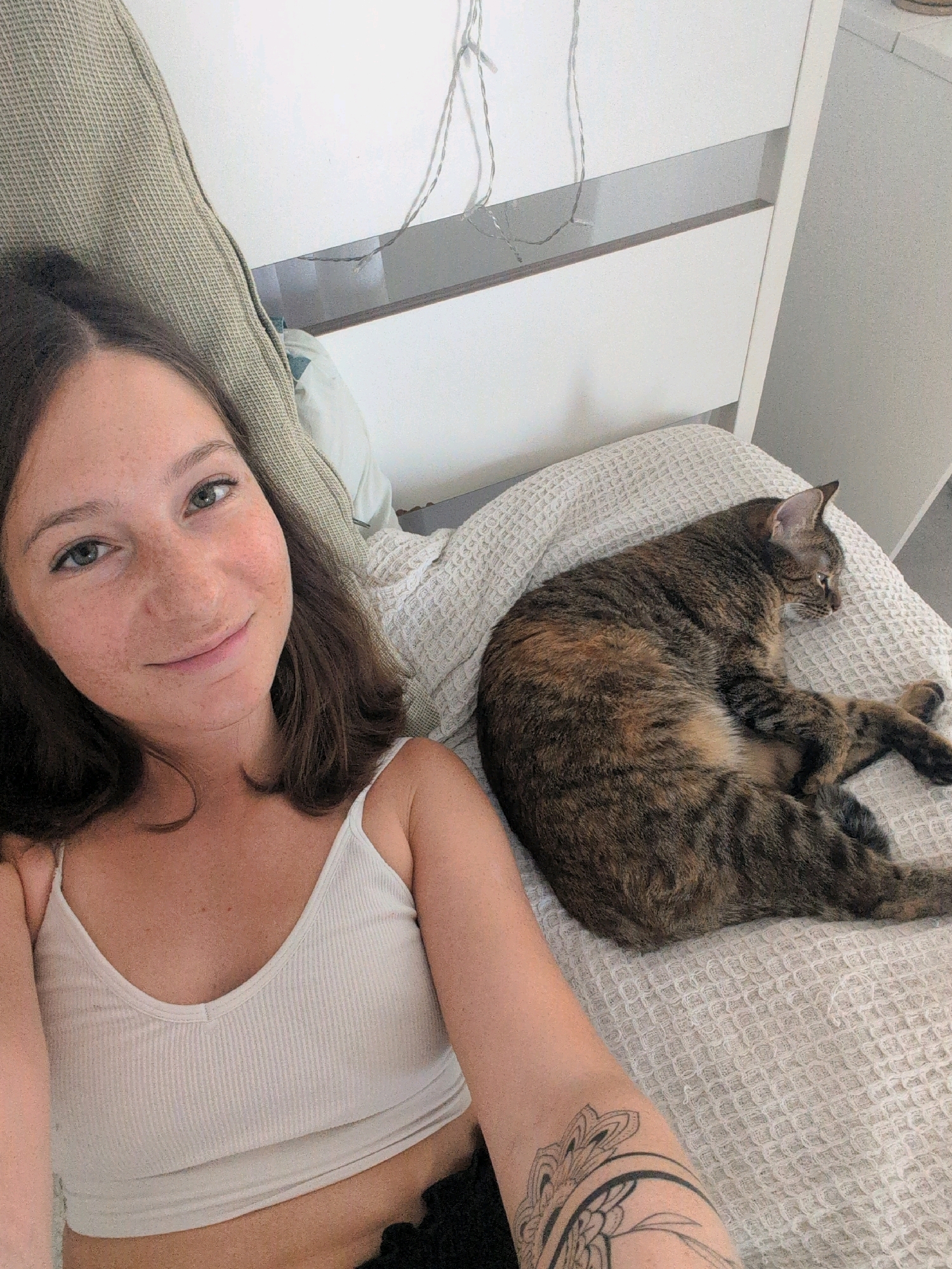 A young woman with freckles and brown hair is lying on a couch with a tabby cat next to her. The woman is wearing a white ribbed tank top and has a tattoo on her arm. The cat is curled up and appears to be sleeping. The couch is covered with a textured blanket. In the background, there is a white dresser with decorative string lights. The scene appears to be indoors.