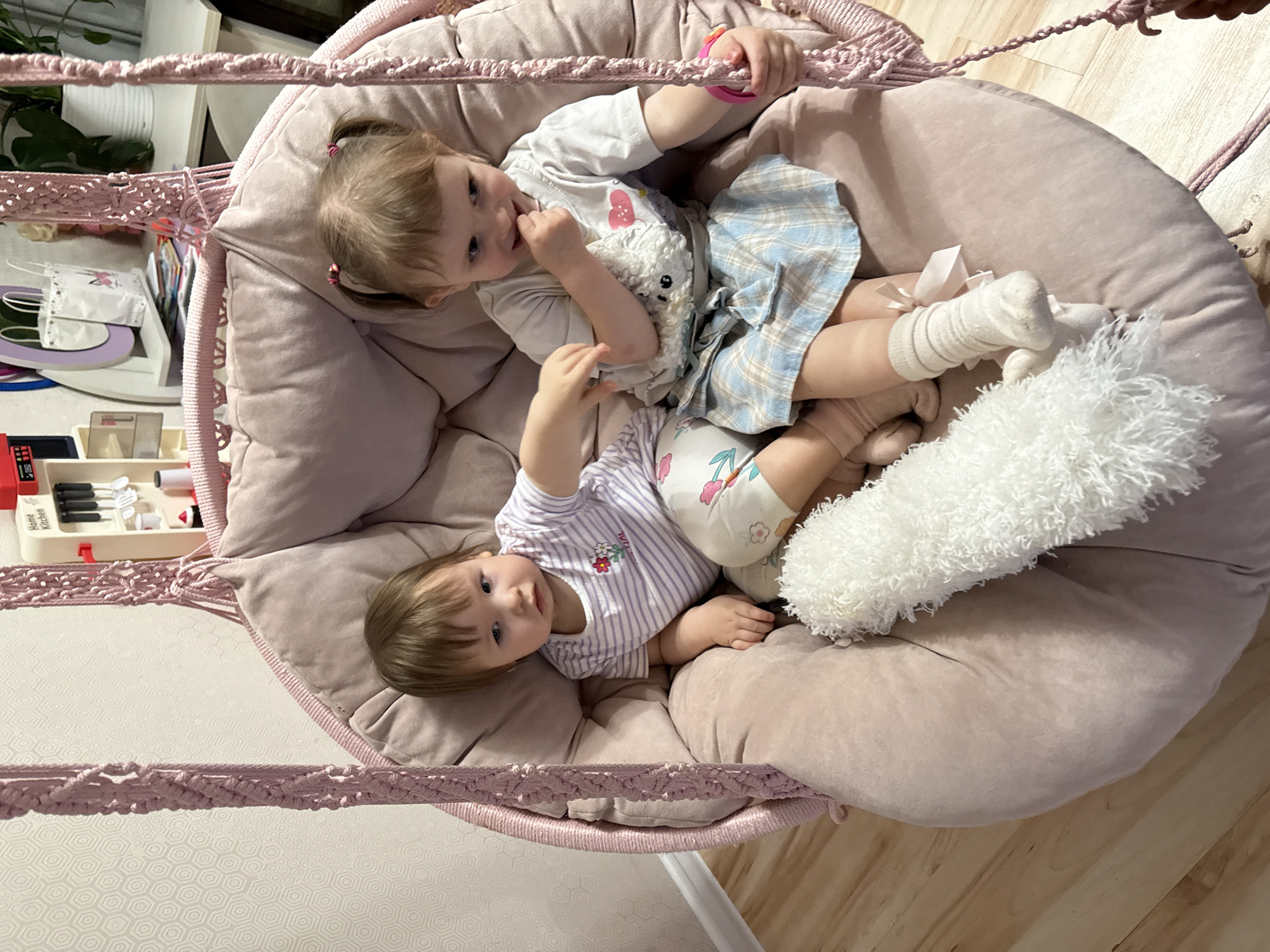 Two young children are lying together in a hanging chair. The chair is round, cushioned, and light pink, suspended by a woven pink cord. The girl on top has blonde hair in pigtails, wearing a white t-shirt, a blue plaid skirt, and white socks. She has a stuffed animal with her. The child below her has short brown hair and is wearing a striped shirt and patterned shorts. There's a white fluffy pillow beside them. The location is indoors, the environment being a domestic residence. There is a child's play kitchen set visible in the background. The image doesn't suggest any location in the city of Krapivka, Russia.