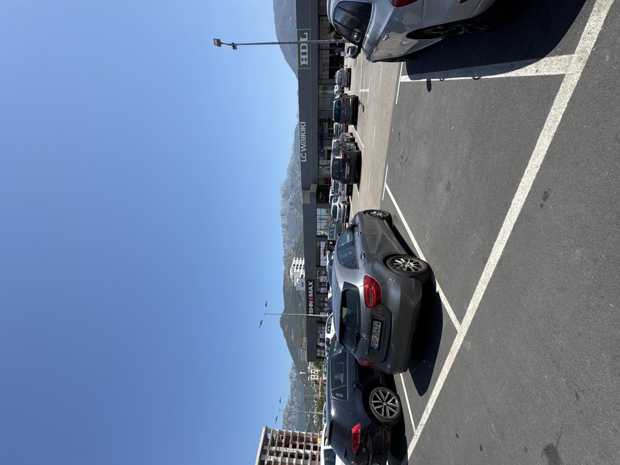 This image depicts a sunlit outdoor parking lot in front of a commercial building, identified as the Cerov Vrh shopping center in Bjeliši, Montenegro. The sky is a clear, bright blue. The parking lot is paved with asphalt and marked with white lines delineating parking spaces. Several cars are parked in the lot, including a dark-colored hatchback on the left and a grey hatchback in the foreground. In the background, a multi-story building under construction is visible to the left, and the main commercial building features prominent signage, including "LC Waikiki" and "HDL". Mountains form the backdrop, suggesting a location near a mountainous region. The overall scene is static, with no individuals or overt actions depicted.