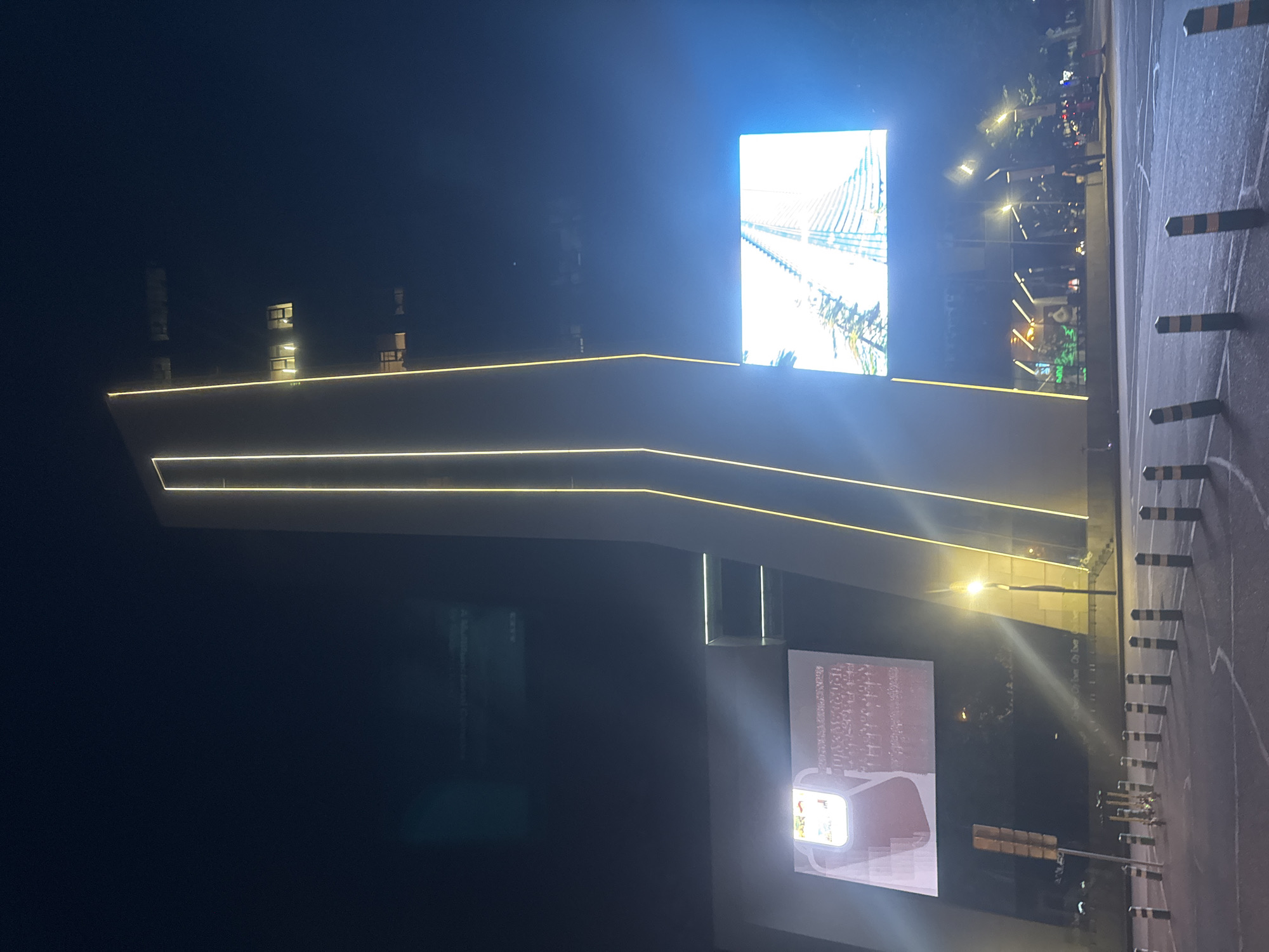 The image depicts a nighttime view of the **Gallery Tbilisi** shopping mall, located in the city of Tbilisi, Georgia. A prominent, illuminated modern architectural structure with bright yellow vertical light strips is the central focus. To the left of this structure, a large digital billboard displays a bright advertisement with images that include a red bag and a pattern of small white squares. To the right, another large digital screen shows a high-angle view of a modern building's facade, appearing white and reflective. The foreground shows a paved area, possibly a street or plaza, with a series of black and yellow bollards delineating a boundary. In the background, the facade of the mall is visible, with some windows illuminated and glimpses of trees and streetlights. A few pedestrians are present in the distance near the mall entrance. The sky is dark, indicating nighttime.