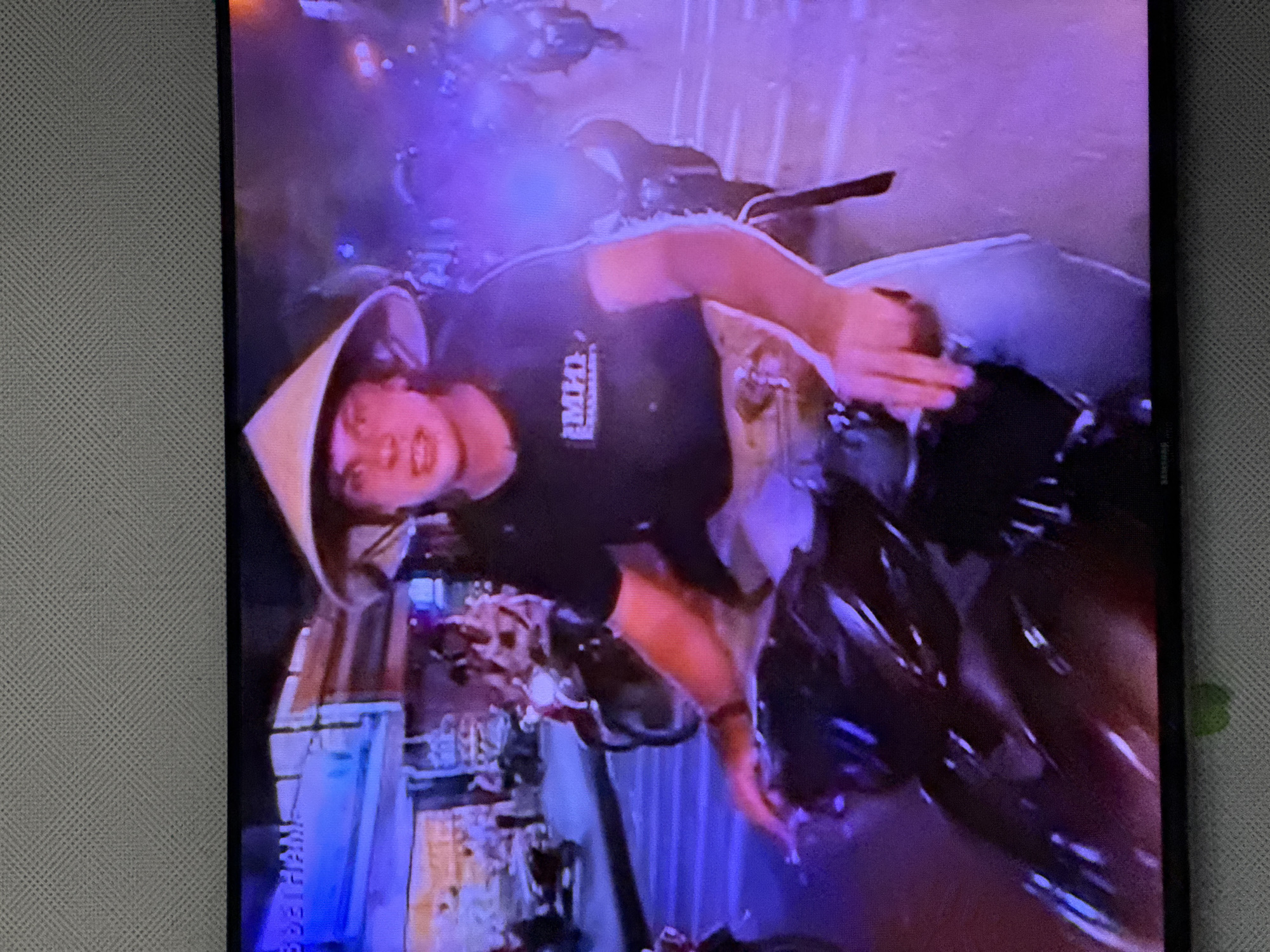 In the foreground, a person wearing a conical straw hat and a black t-shirt with a white logo is riding a motorcycle. The person's face is visible, and they appear to be looking at the camera. The t-shirt logo appears to be rectangular with text inside. The person is wearing light-colored pants. The motorcycle is dark-colored. In the background, other motorcycles and people are visible, as well as buildings and streetlights. The scene appears to be a street in a city at night, with purple and blue lighting dominating the image. The overall impression is that of a busy urban environment at night. The location is the city of Oskemen, Kazakhstan.