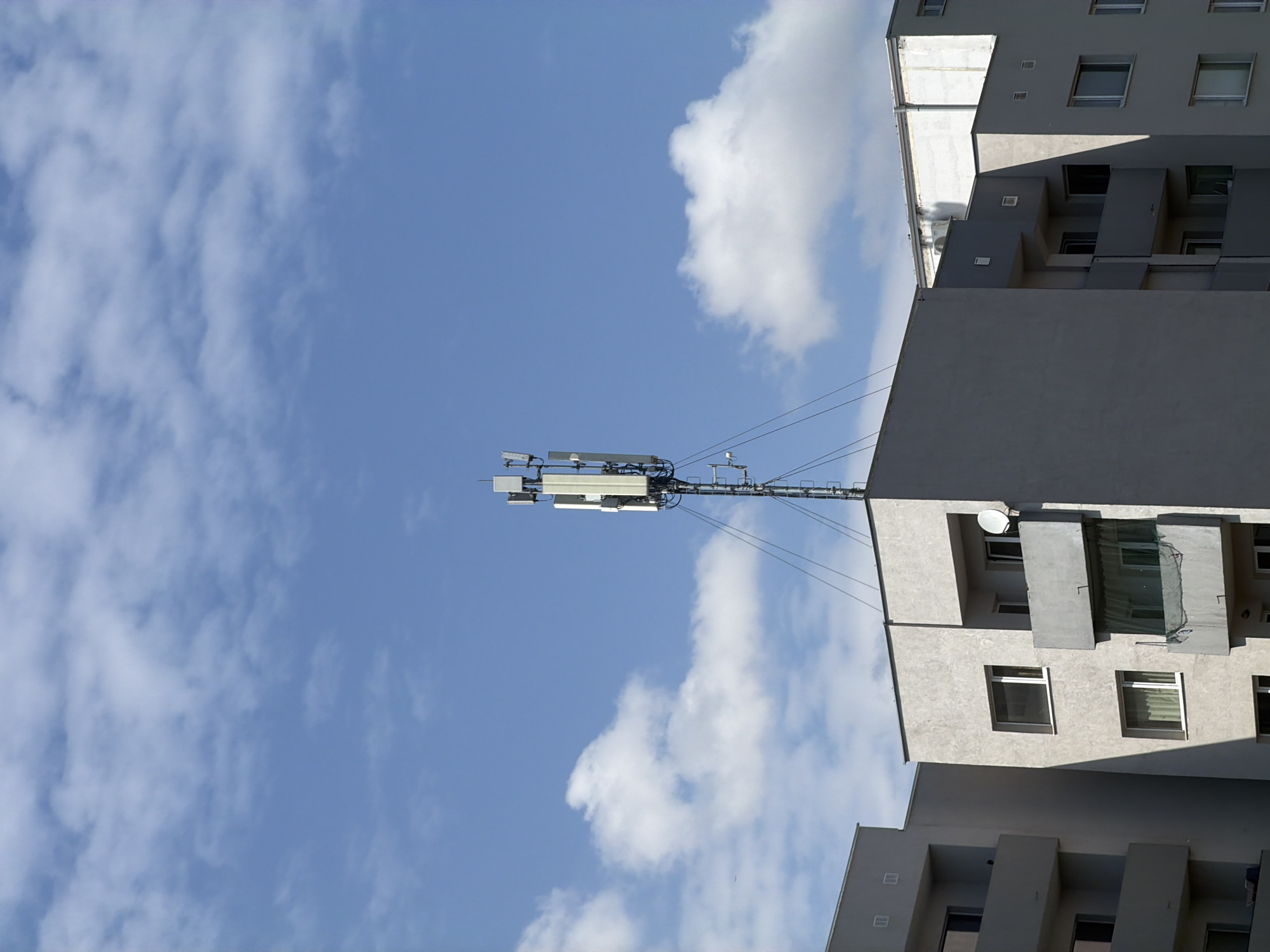 The image shows a building in Sofia, Bulgaria, with a cell tower affixed to its roof. The building is of modern construction, featuring a grey facade and cubist architectural elements. The sky is visible behind the building, with white clouds against a blue sky. The cell tower has multiple antennae and supporting structures, and is wired to the building.