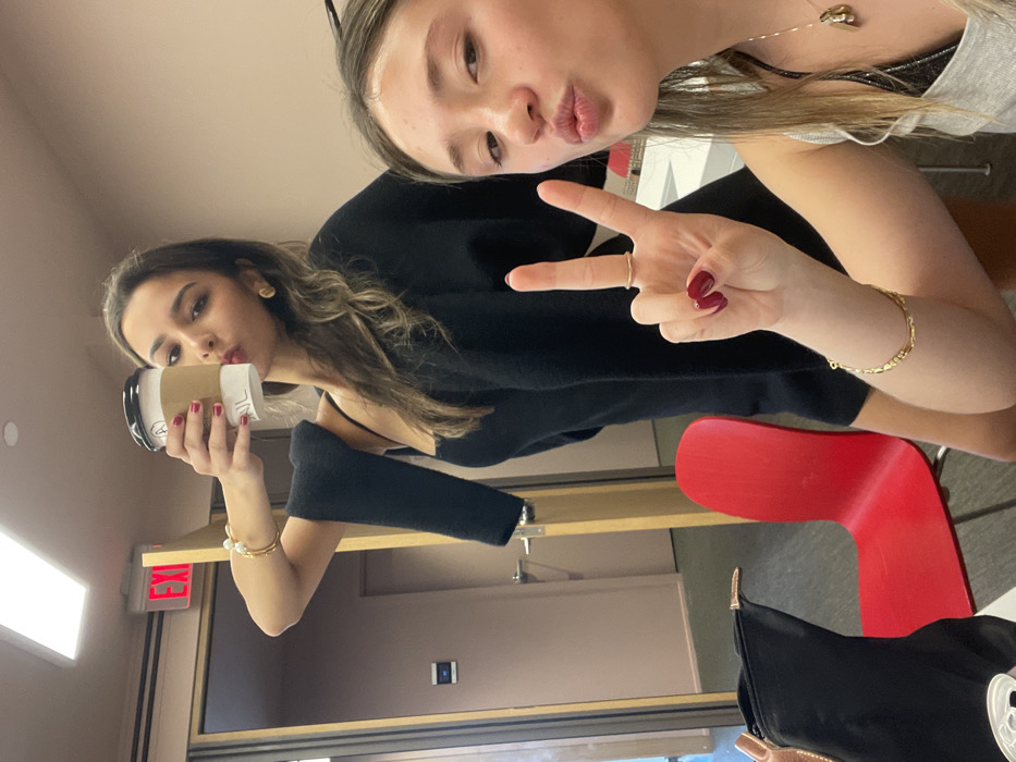 Two young women pose closely for a selfie in what appears to be an indoor space. One woman, partially visible, makes a peace sign. The other holds a coffee cup and pouts her lips. Both have dark manicured nails, and are wearing jewelry. The setting includes a red chair, a black purse, and a door with an 'EXIT' sign visible above.