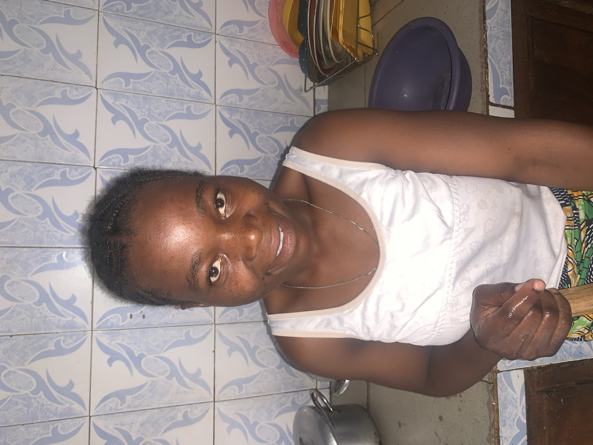 A medium shot depicts a dark-skinned woman indoors. She is wearing a white tank top and a thin silver necklace. Her hair is short and dark. She is smiling and looking directly at the camera. Her hand appears wet and slightly clenched. A cooking pot is visible in the lower left corner. The background includes blue and white patterned tiles, a dish rack with colorful plates, a purple bowl, and a section of a dark wooden door. There is no verifiable information in the image to confirm the specific location as Jos, Nigeria. The presence of cooking utensils and patterned tiles indicates the environment is likely a kitchen.