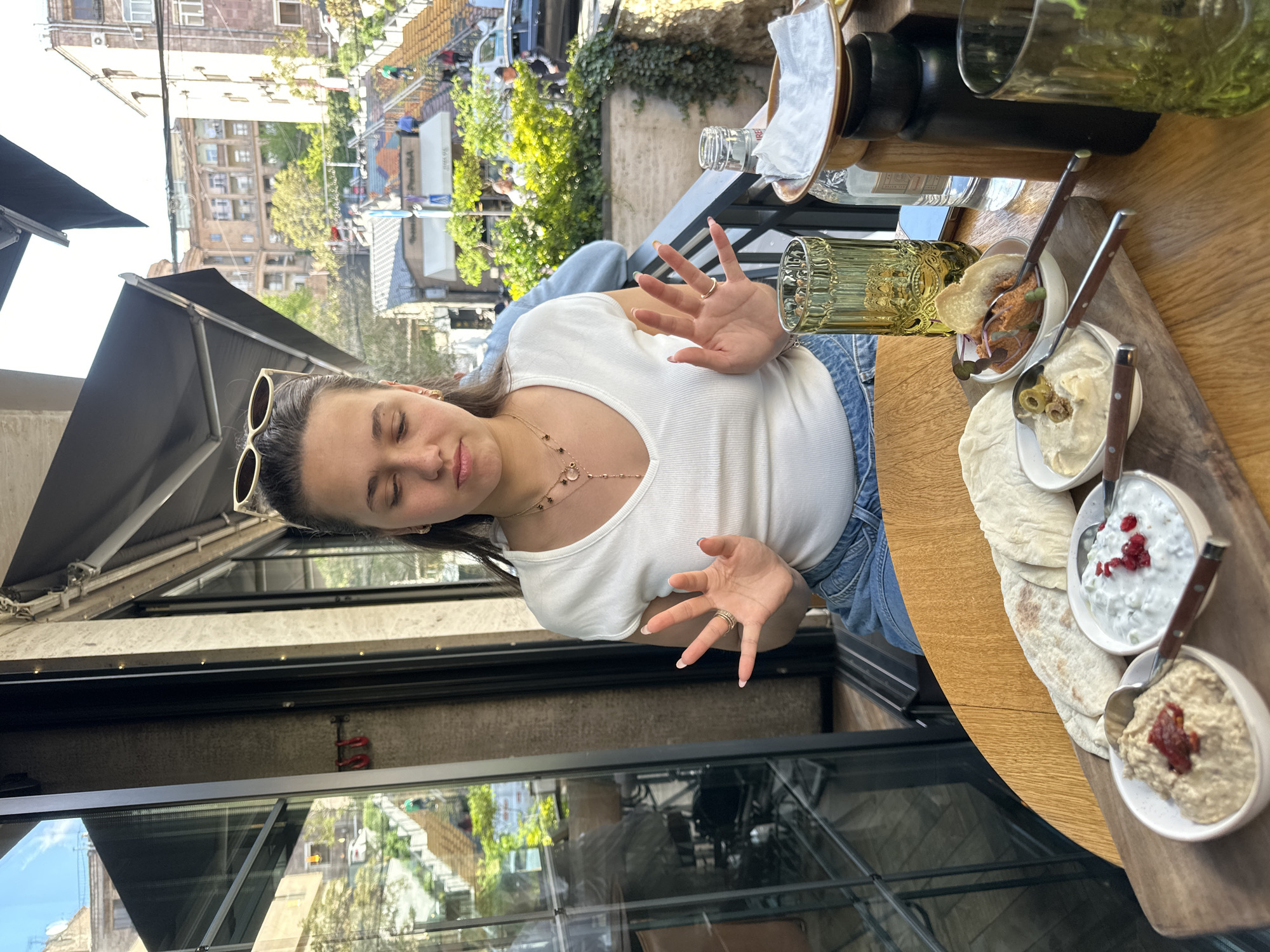 A young woman is captured in a medium, tilted shot at a restaurant, likely in Yerevan, Armenia, based on architectural styles. She wears a white v-neck top, blue jeans, and gold-rimmed sunglasses perched on her head. A delicate gold necklace adorns her neck. Her eyes are closed, and she extends both hands in front of her.
The scene is set on a restaurant balcony with a wooden table. On the table is a wooden board with several small white bowls containing various dips. Flatbread is arranged between the bowls. Glassware, including a textured green glass and a water bottle, is also visible.
The restaurant's exterior shows black-framed glass windows and a black awning. Buildings and trees can be seen in the background across the street. A car is also parked on the street. The lighting suggests it is daytime.