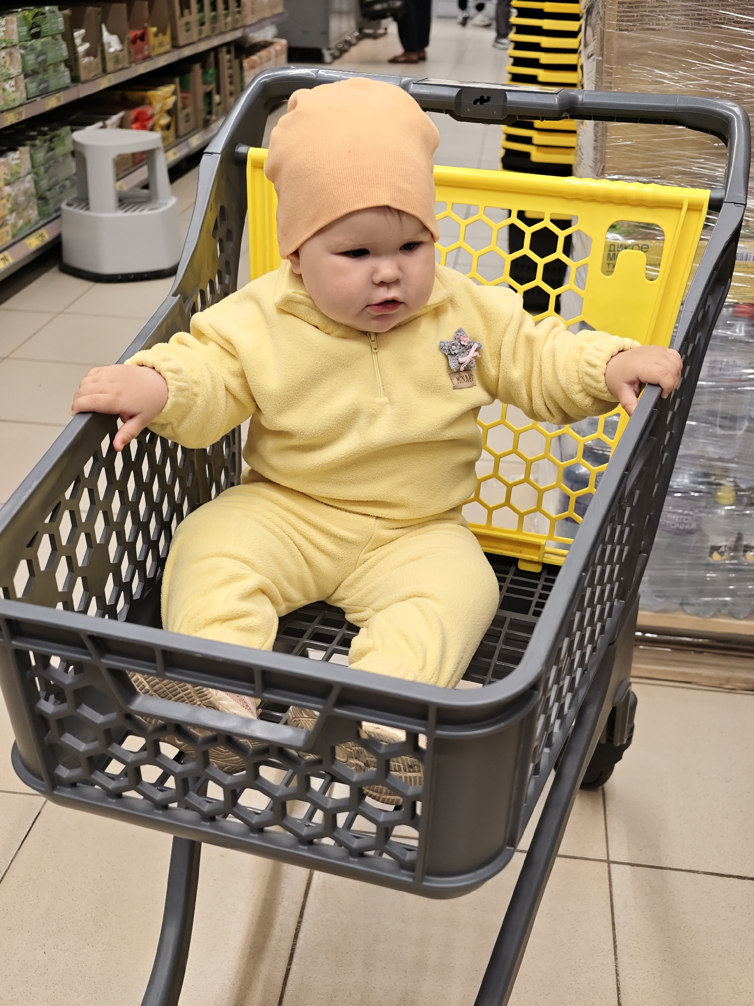 A baby wearing a yellow outfit and a peach-colored hat sits in a gray shopping cart, which has a yellow divider. The cart is in a grocery store aisle, with shelves stocked with products visible in the background. The floor is tiled. The scene is likely in a store in the city of Chaikovsky, Russia.