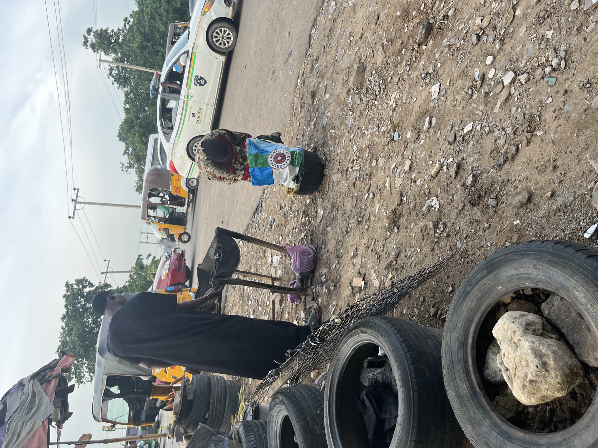 The image depicts an outdoor scene on the side of a road in Abuja, Nigeria. In the foreground, several discarded tires are piled up. A person wearing a black traditional outfit is standing next to a makeshift grill or brazier. Further to the right, another person is bent over, interacting with a large bag of "Golden Penny Foods" and a black basin, which appears to contain corn. In the background, various vehicles, including a yellow and brown auto-rickshaw (Keke Napep), a red car, and a white police vehicle with "NIGERIA POLICE" and "RAPID RESPONSE" markings, are present on the road, indicating traffic. Trees line the distant horizon, and the sky is overcast. The ground is a mixture of dirt, gravel, and debris. The overall impression is of a busy roadside area, possibly a marketplace or a place where street vendors operate.