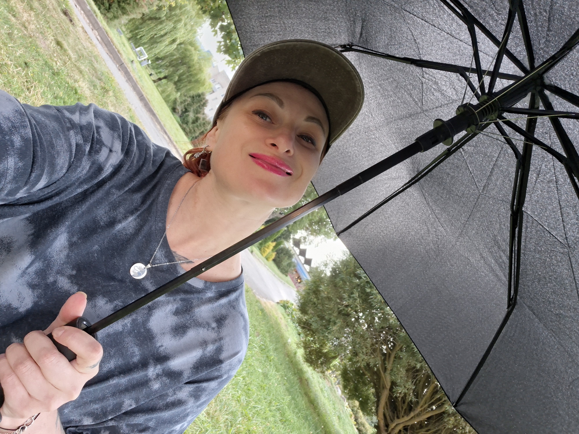 A woman is pictured from the chest up, holding an open black umbrella over her head. She wears a dark grey tie-dye t-shirt, a green baseball cap, and a silver necklace with a circular pendant. She is looking up and to her left, smiling, with bright pink lipstick. Her right hand is visible, gripping the umbrella handle. The background shows a grassy slope with a paved path and trees. The scene is outdoors, likely in a park or residential area, with buildings faintly visible in the distance. The overall impression is of a casual, perhaps slightly humid day.