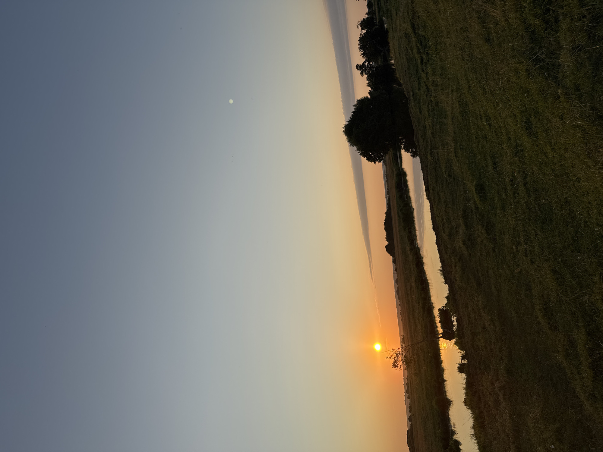 The image depicts a rural landscape at sunset in Boriskino, Russia. The sky transitions from a pale blue at the top to warm orange hues near the horizon. The sun, a bright orange orb, is setting on the left side of the frame, casting a golden reflection on a calm river that winds through a grassy field. The riverbanks are lined with vegetation and a few scattered trees. On the right side of the river, a cluster of darker trees stands in silhouette against the fading light. In the far distance, a faint outline of hills or a treeline is visible. There are no people or animals visible in the scene. The overall impression is one of tranquility and natural beauty as day transitions into night.