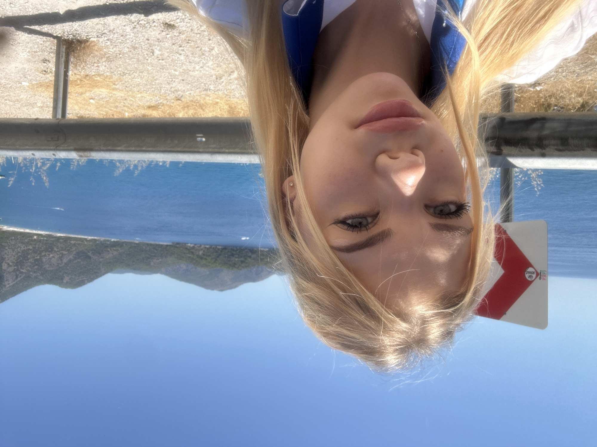 A selfie taken outdoors shows a young woman with blonde hair and light-colored eyes, her face close to the camera. She is wearing a white shirt with blue trim and a necklace. Behind her, a bright blue body of water is visible, bordered by a coastline with green, tree-covered hills. A traffic sign with a red and white arrow shape is to her left, and a metal guardrail runs horizontally across the middle ground. The sky is clear and blue.
