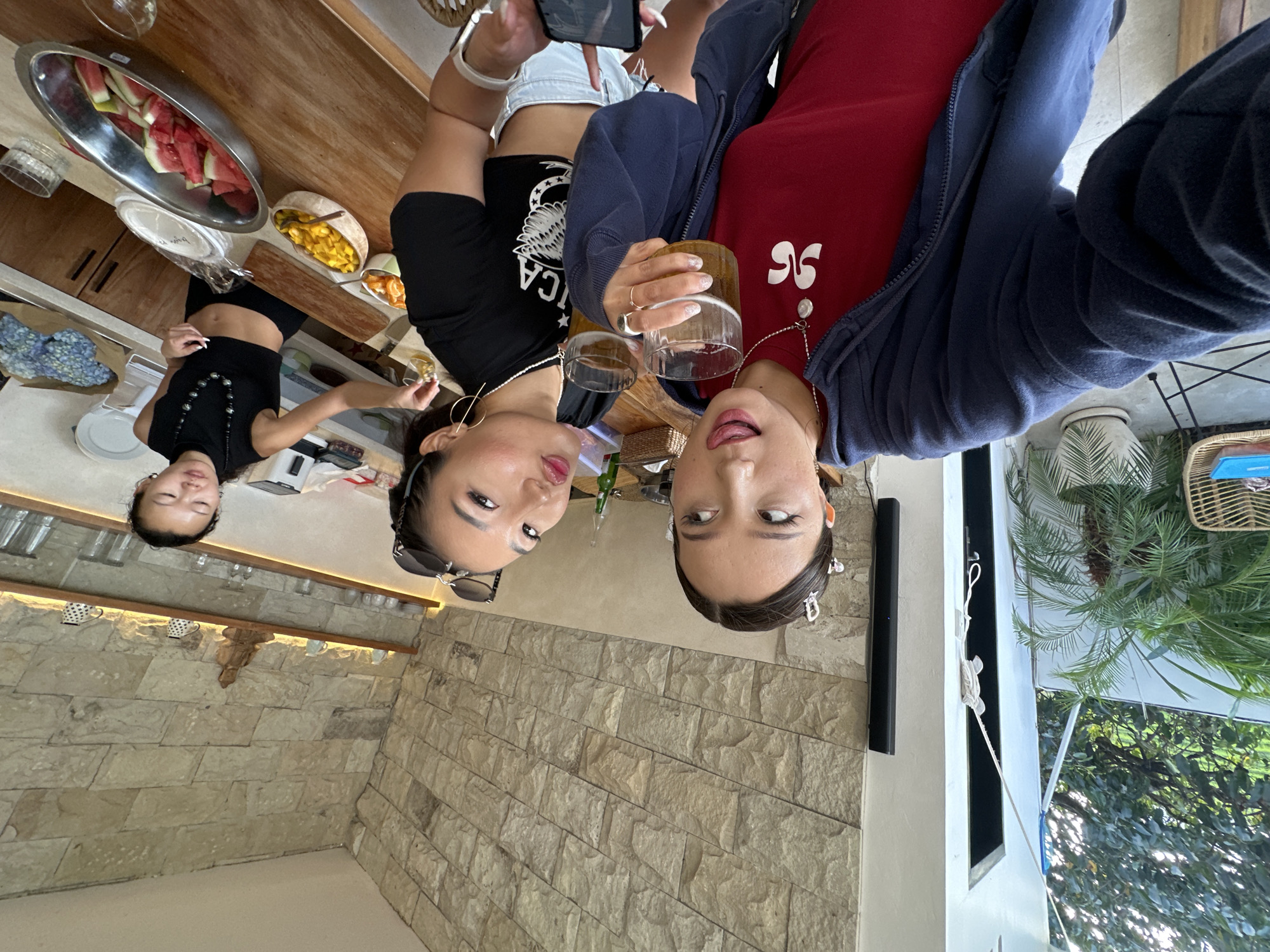 The image depicts an indoor-outdoor scene, likely a common area such as a kitchen or dining space, located in Mengwi, Indonesia. Three East Asian females are present.
The primary focus is on two females in the foreground, positioned for a selfie. The female on the left, holding the camera, wears a dark blue zipped hoodie over a red t-shirt with a white stylized "96" emblem and a silver necklace with a white pendant. Her dark hair is pulled back with multiple decorative clips, and she has heavy eyeliner. She holds a glass of amber liquid (possibly beer) in her right hand, extends her tongue slightly, and looks upwards to the right. The female beside her, to the right, wears a black cropped t-shirt with a partial white "AMERICA" and eagle graphic, light-colored shorts, and gold hoop earrings. Sunglasses rest on her dark, pulled-back hair. She holds a similar glass of amber liquid in her left hand and puckers her lips towards the camera. Her right hand, with manicured nails, holds a smartphone pointed downwards.
Behind the two selfie-takers, the room features a textured wall composed of light-colored, irregularly shaped stone tiles, extending from the mid-ground to the ceiling. Below this wall, a long, thick wooden counter or bar runs across the mid-ground. Above the counter, two illuminated wooden shelves display various clear glasses, white cups, and a small wooden Buddha statue in the center of the top shelf.
On the wooden counter, from left to right, are a small bowl of orange fruit segments, a boat-shaped wooden bowl containing diced yellow fruit (mango), and a larger oval metal bowl filled with sliced red and green fruit (watermelon). A stack of white paper plates is visible in a plastic bag. Further to the right, a third female stands behind the counter, appearing to prepare or serve items. She wears a black high-necked sleeveless crop top and a black beaded necklace. Her right hand holds a small glass item over a clear dish. A white appliance, possibly a blender or food processor, is also visible on the counter. A blue floral fabric item rests on the far right of the counter.
To the left of the selfie-takers, an open doorway or archway provides a view to an outdoor area with lush green foliage, including palm plants, and a light-colored wicker chair. A white braided rope-like decoration hangs down vertically near the doorway.