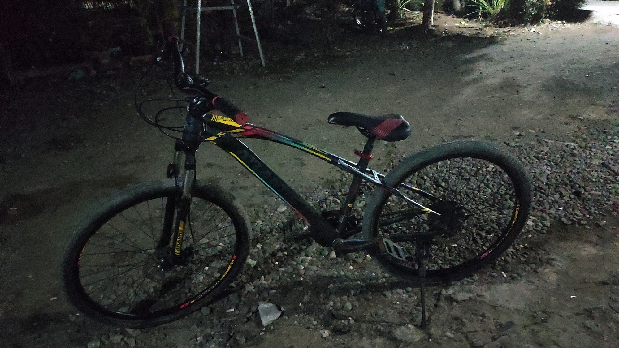 A black mountain bike with multi-colored accents on the frame is parked on a gravel surface at night. The bicycle has a black seat with red accents, black handlebars, and black tires with yellow rims. A kickstand is deployed, supporting the bike. In the background, a dimly lit outdoor area is visible, with shadows and some foliage. The overall scene suggests a residential or informal outdoor space in Tanjungan, Indonesia.