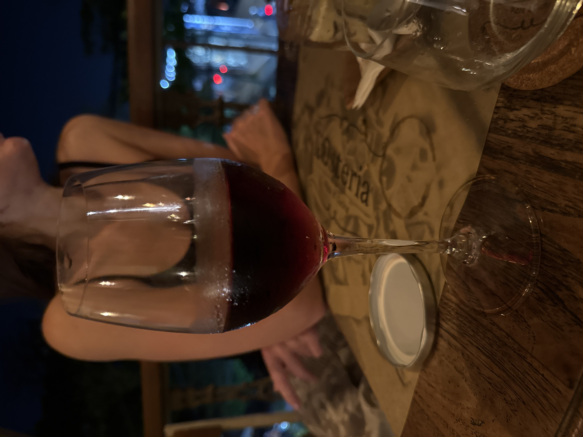 The image shows a wine glass filled with red wine on a wooden table. A white cap rests under the stem of the glass. In the background, a blurred cityscape with bokeh lights is visible. The image includes a partial view of a person's face and upper body, appearing to be at a restaurant or outdoor venue. The table setting includes a paper placemat with some text, partially obscured, likely the restaurant's name "Osteria".
