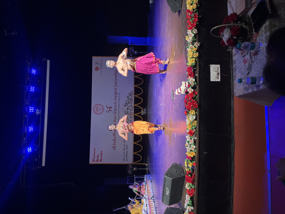 Two men in traditional Indian clothing are performing a dance on a stage. They are both wearing bright colored costumes and are holding their arms in the air. The stage is decorated with flowers and the banner behind them says "54th All India Bhar Rao Nritya & Sangeet Sammelan" and "Celebrating the Soul of Indian Classical Arts". The stage is lit with bright lights and the audience is seated in the dark. The location is the Pracheen Kala Kendra, Sector 35, Chandigarh, India, with the coordinates (latitude: 30.733, longitude: 76.789). There is a microphone and a speaker on the stage to the left of the performers, and a basket of flowers is on the table in the foreground.