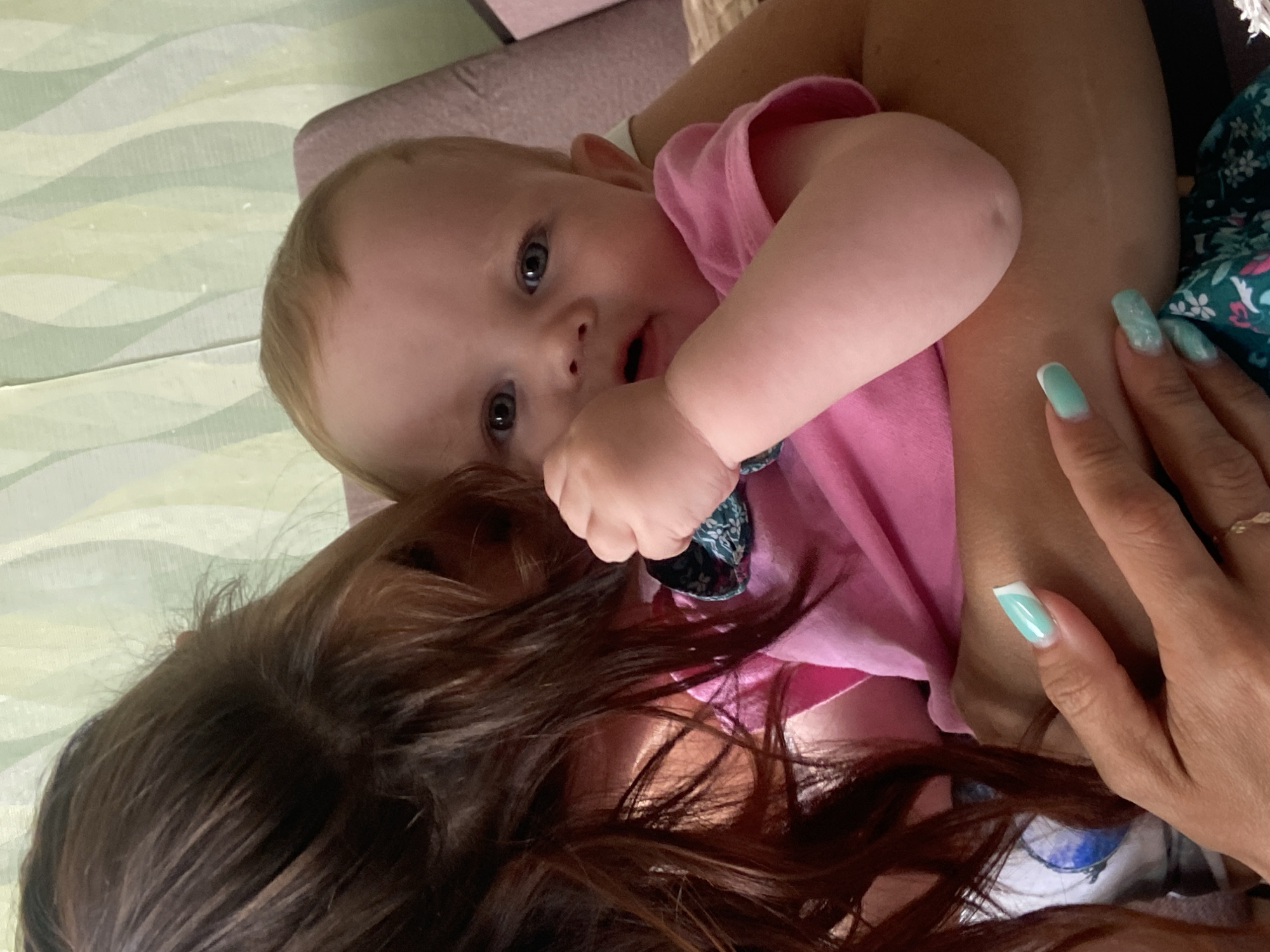 A close-up, angled shot shows a baby with light skin, blue eyes, and blond hair wearing a pink shirt, being held. Part of an adult with brown hair and manicured fingernails with light blue polish is visible. The location is unknown, but possibly in a residence. The baby is looking at the camera. A green and dark floral patterned scrunchie is around the baby's wrist. The background is a patterned wall and a soft pink sofa. It is daytime, indoors.