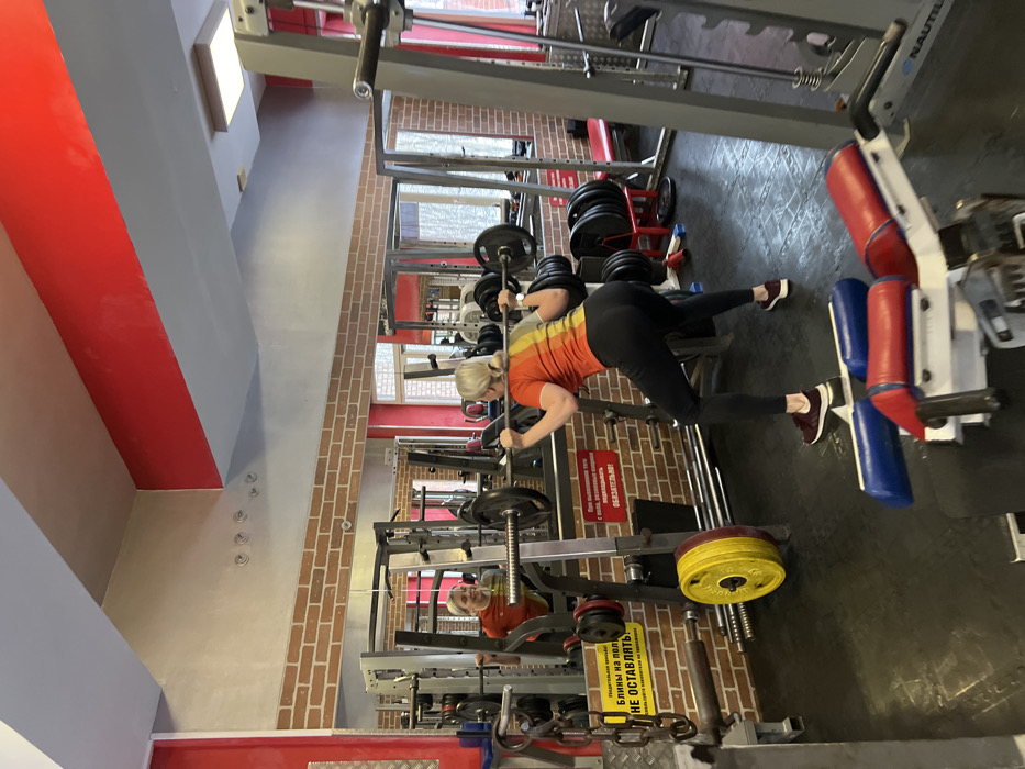 A woman is working out in a gym. She is wearing a bright orange and yellow striped shirt, black pants, and maroon sneakers. She has a blonde ponytail. She is positioned in a squat rack, lifting a barbell loaded with weights. Other weight machines and gym equipment are visible in the background. The walls are brick and painted red and grey. A red and white warning sign is near her, and another person is reflected in a mirror to the left.