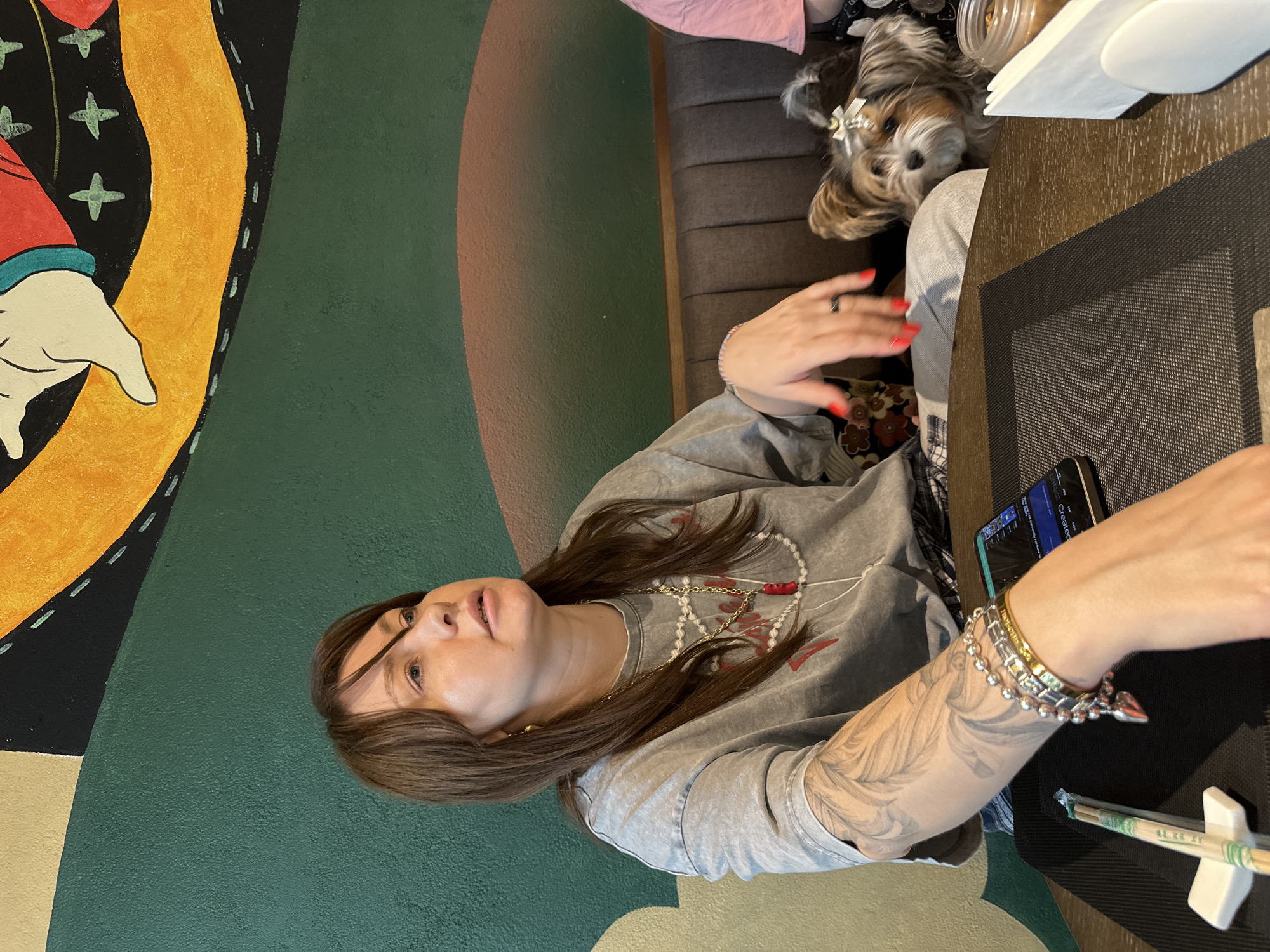 The image captures a casual scene inside what appears to be a restaurant or cafe in Moscow, Russia. The primary subject is a woman with long, brown hair, wearing a grey t-shirt, a long pearl necklace, and a gold chain. She has an intricate grey and black tattoo on her right forearm, featuring flowing, feather-like patterns, and multiple bracelets on her wrist. Her fingernails are painted red. She looks slightly upwards and to her right with a calm, somewhat contemplative expression.
Seated behind her, on a dark grey upholstered booth, is a small, fluffy dog, likely a Yorkshire Terrier, with brown, grey, and white fur, adorned with a white bow in its hair. The dog looks alertly towards the left side of the frame.
In front of the woman is a dark brown, textured table with a black woven placemat. Green and white chopsticks are resting in a white holder on the placemat, suggesting a meal, possibly East Asian cuisine. A smartphone or small tablet with a lit screen, partially displaying the word "Created," is also on the table. Behind it, a small glass jar containing a light brown substance (possibly sugar or spices) is visible, topped with a white napkin.
The background features a vibrant wall mural with abstract shapes and bold colors including dark green, yellow, black, red, and off-white, adding a dynamic artistic element to the setting. The overall impression is a relaxed indoor dining experience, possibly in a pet-friendly establishment.