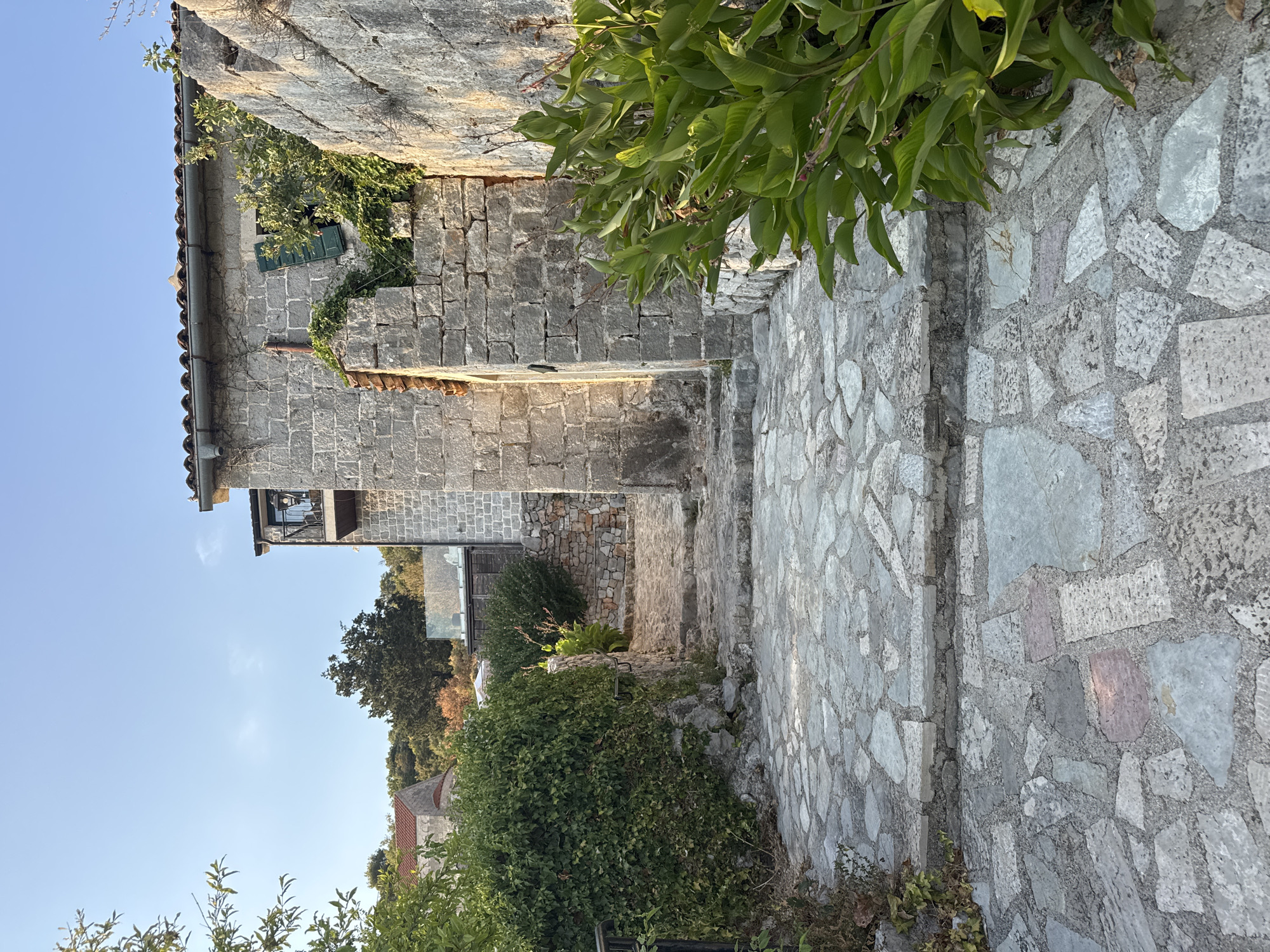 The image shows a stone pathway leading up a gentle incline. The pathway is made of irregularly shaped flagstones in various shades of gray and some with reddish hues. To the left, a thick hedge of ivy covers a stone wall and a portion of the pathway. To the right, a large stone wall runs along the pathway, and large green leafy plants grow in front of it. In the background, a stone building with a tiled roof and a balcony is visible. The sky is a clear blue with a few faint clouds. The scene appears to be in a historic urban setting, possibly a Mediterranean town.
