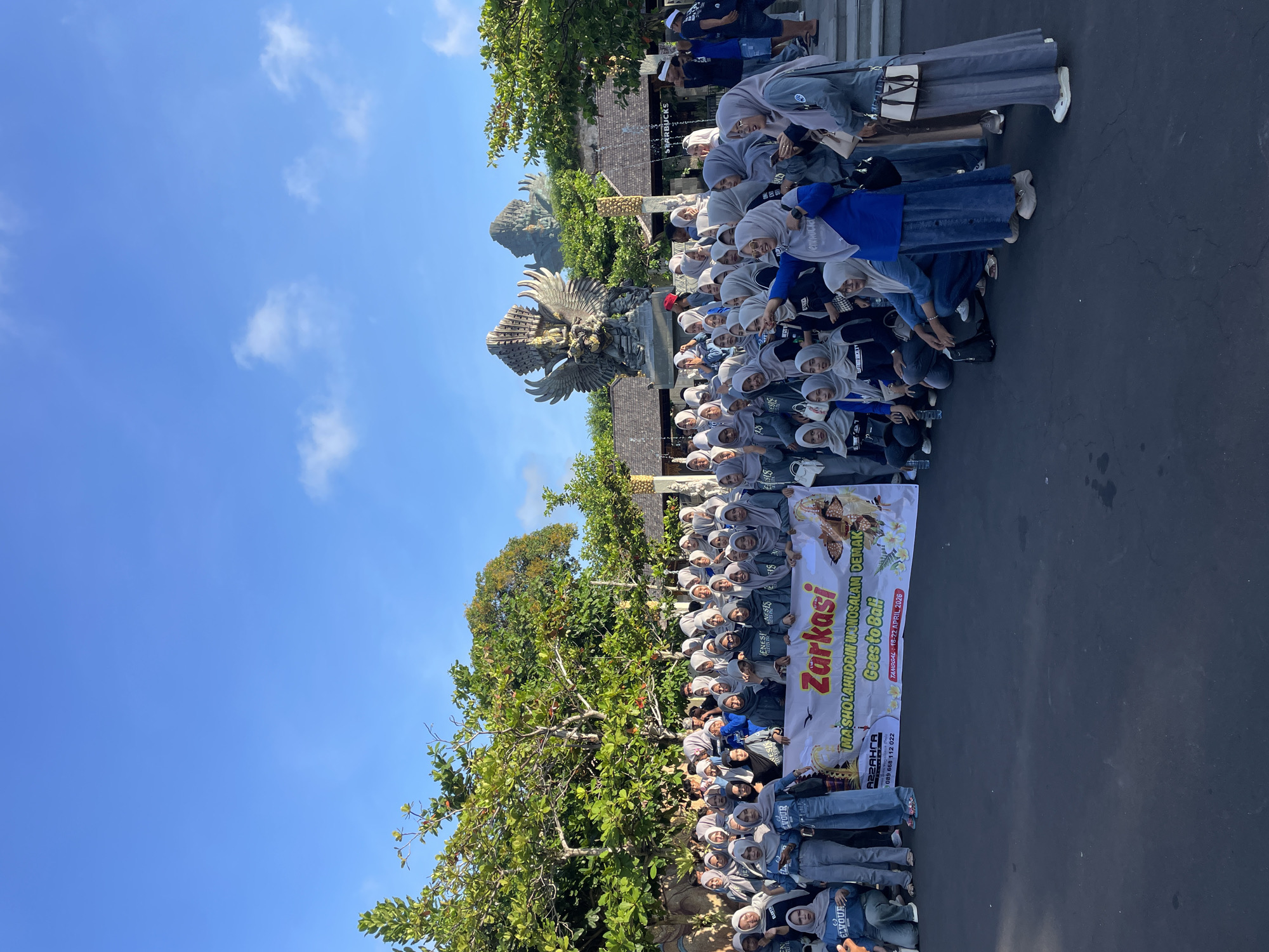 A large group of people, mostly women wearing hijabs, are standing in a formation and posing for a photograph in front of a statue in Kuta Selatan, Indonesia. They are holding a banner that reads "Zarkasi MA SHOLAHUDDIN WONOSALAM DEMAK Goes to Bali TANGGAL: 18-22 APRIL 2026." The statue appears to be Garuda Wisnu Kencana. In the background, there is a Starbucks sign. The sky is blue and clear.