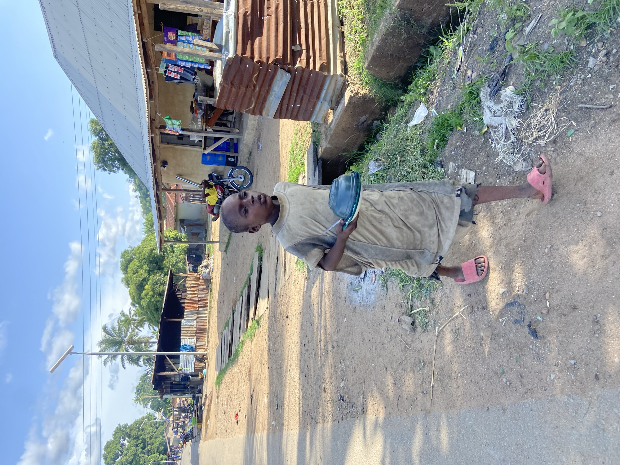 A boy wearing a light-colored shirt, shorts, and pink sandals stands on a dirt shoulder alongside a paved road in Mararaba, Nigeria. He holds a blue pan and tool. Behind him, a corrugated metal structure with a variety of goods for sale. A motorcycle and other buildings are visible along the road, and a street light. Debris and vegetation are along the side of the road. A cement drainage ditch with grass growing up from it is visible on the right. The sky is blue with scattered clouds.