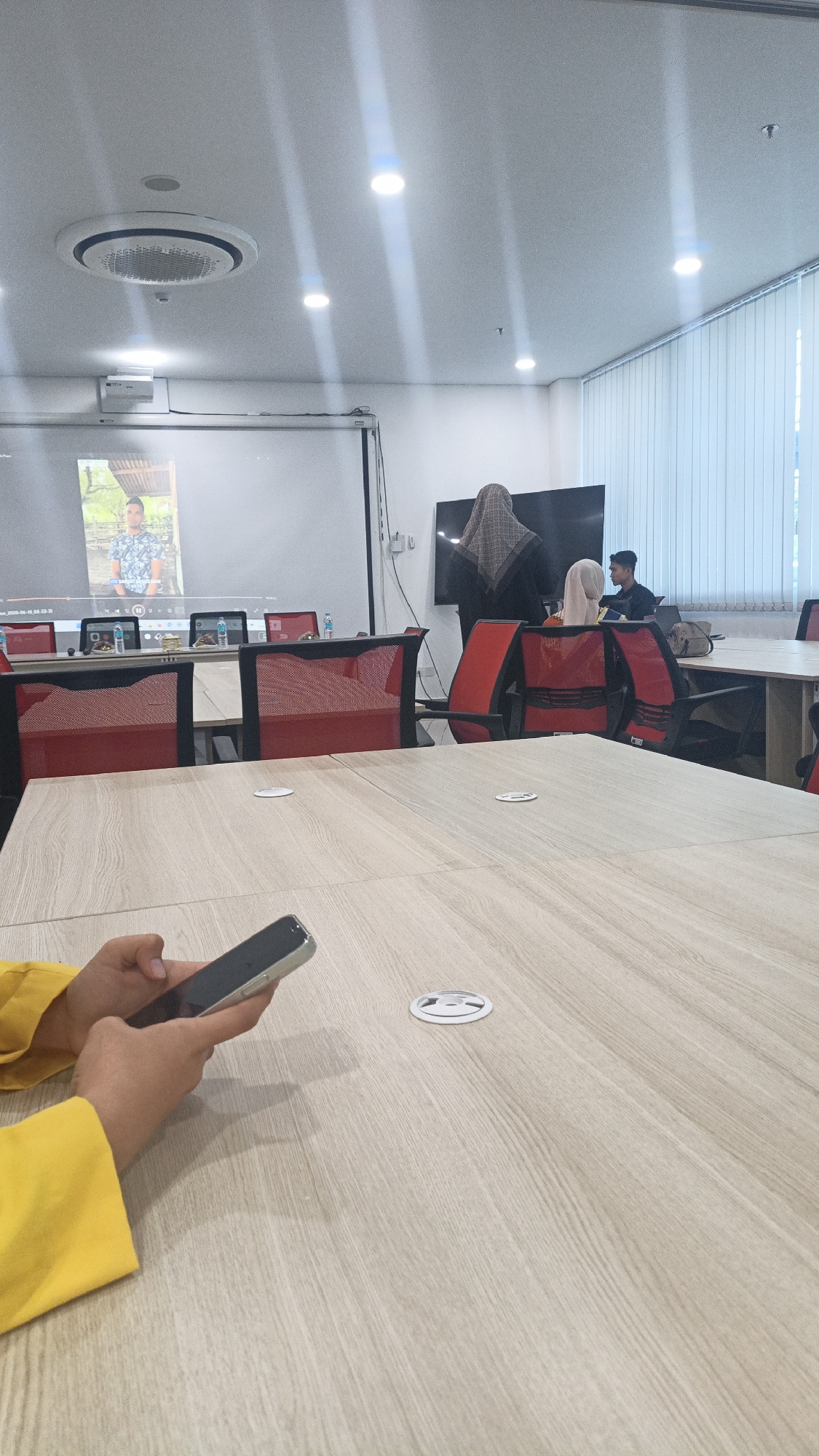 The image depicts a meeting room in Lhokseumawe, Indonesia. A group of people are gathered around a conference table. In the foreground, a person's hands in a yellow top are holding a smartphone. The background shows a large projector screen displaying an image of a man in a patterned shirt. Further back, two women wearing hijabs are standing, and a young man is seated at a laptop.
The room is equipped with a projector, a large screen, and a conference table with red chairs. The lighting suggests it is daytime, with natural light filtering through blinds on the right side of the room. The overall atmosphere appears to be a professional meeting or presentation. There is no visible text in the image that provides specific details about the event or time.