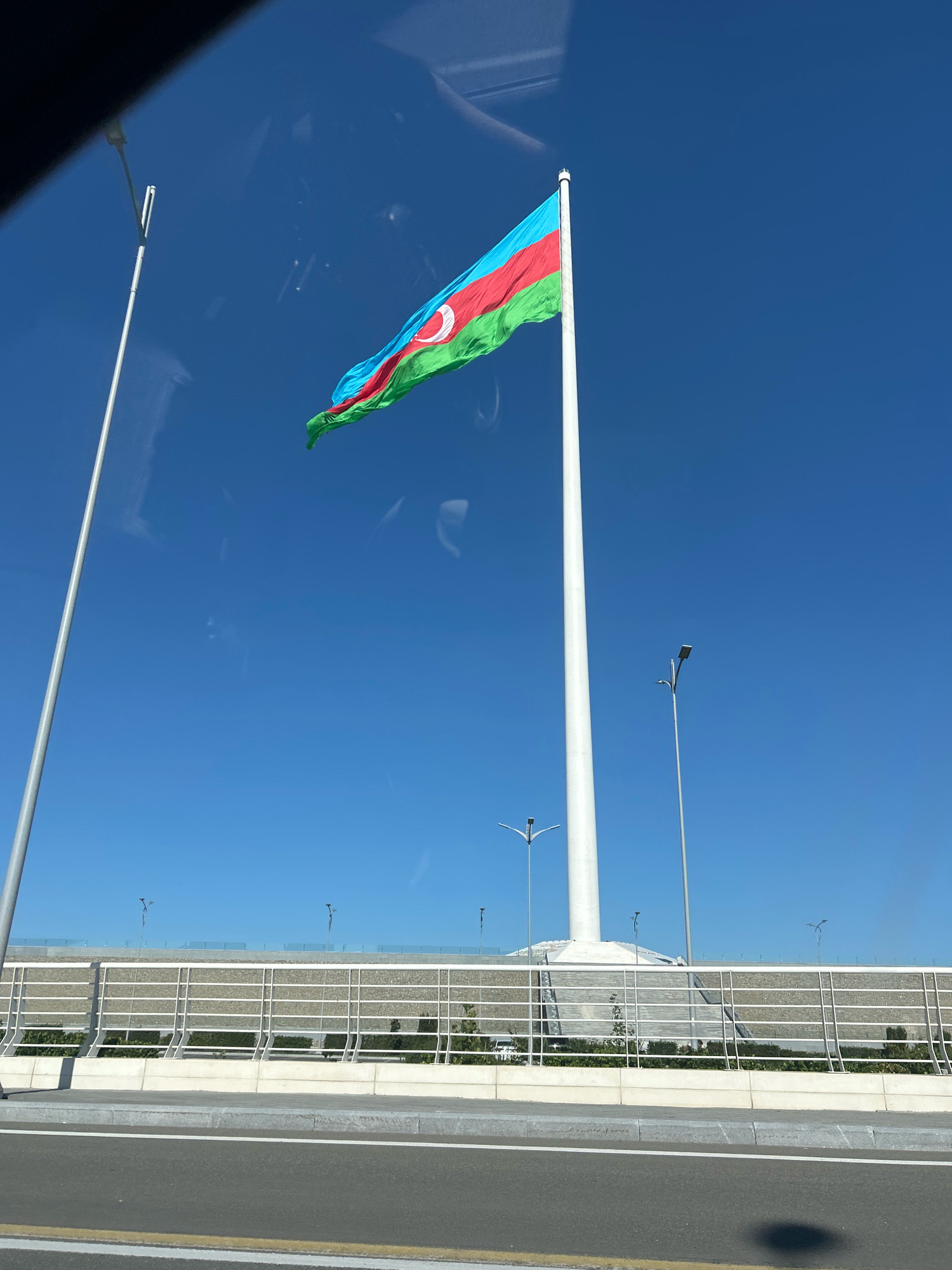 The image shows a wide, white flagpole with the flag of Azerbaijan flying from it, against a clear blue sky. The flag is composed of three horizontal stripes of blue, red, and green, with a white crescent and star in the center of the red stripe. The flagpole is located on the right side of the image, with a large concrete base visible at its bottom. In the background, there is a multi-tiered structure with white railings and some greenery. The foreground shows a road with white and yellow lines. The scene appears to be an outdoor area, possibly a park or a public square. The image was captured in the Sabail Raion of Azerbaijan.