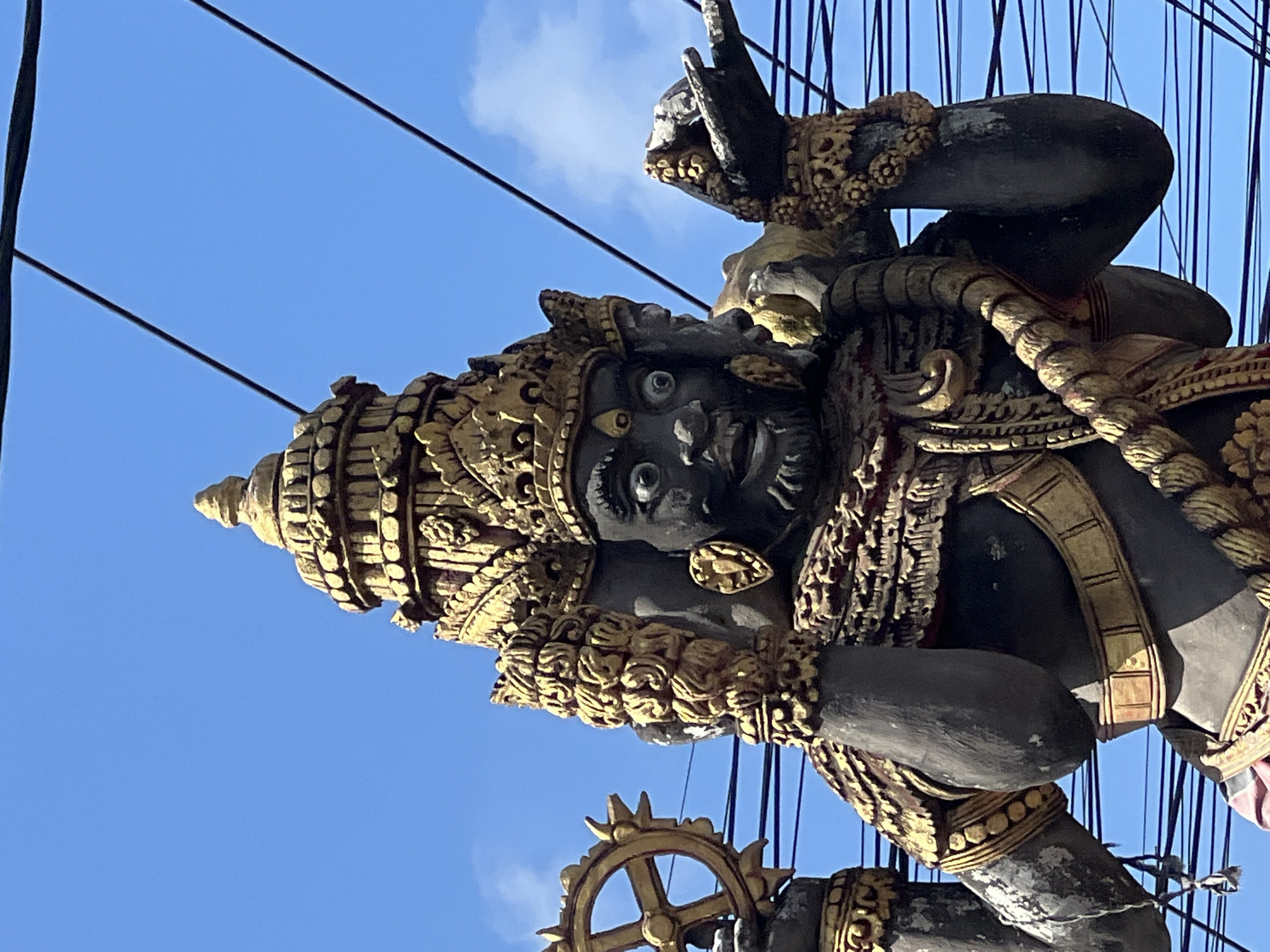 A statue of a multi-faced deity, possibly Hindu, stands against a bright blue sky with scattered clouds. The statue is dark-colored with intricate gold ornamentation. It appears to have at least four faces, with the central face prominently displaying wide, startled eyes and an open mouth. The deity is adorned with elaborate gold crowns, necklaces, armbands, and a sash. In its left hand, it holds a golden chakra or wheel, and its right hand is raised, palm open, with an object that appears to be a skull or a similar macabre item. The statue is positioned outdoors, with numerous dark electrical wires crisscrossing the frame in front of it. The location is identified as North Kuta, Indonesia.